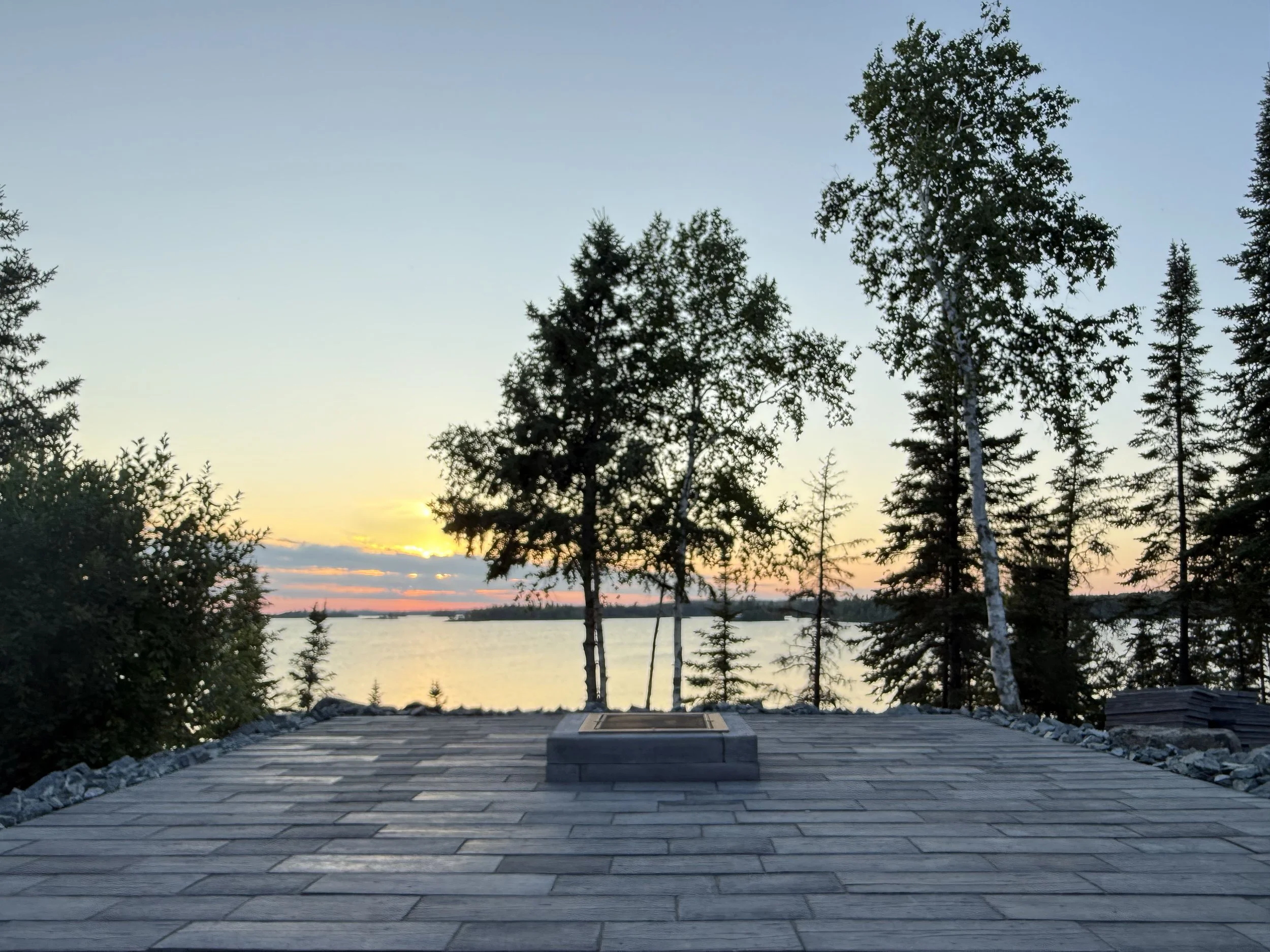 Sunset over a lake with trees in the foreground and a paved viewing platform in the front.