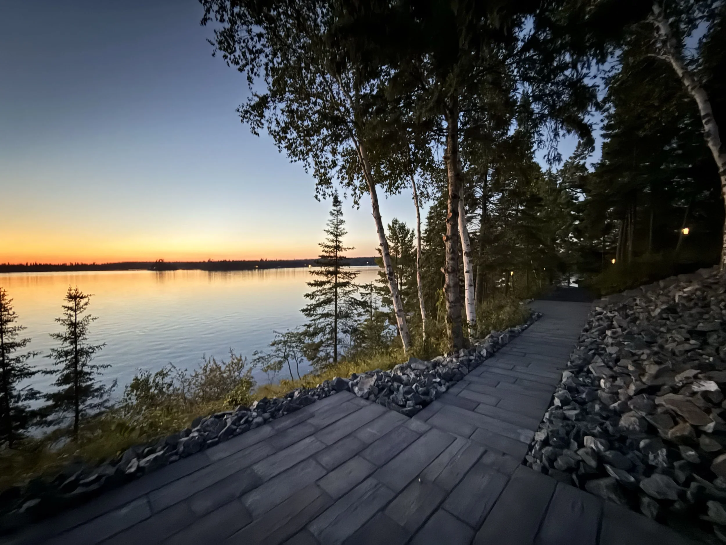 Sunset over a calm lake with a rocky shoreline and a wooden walkway, surrounded by trees.