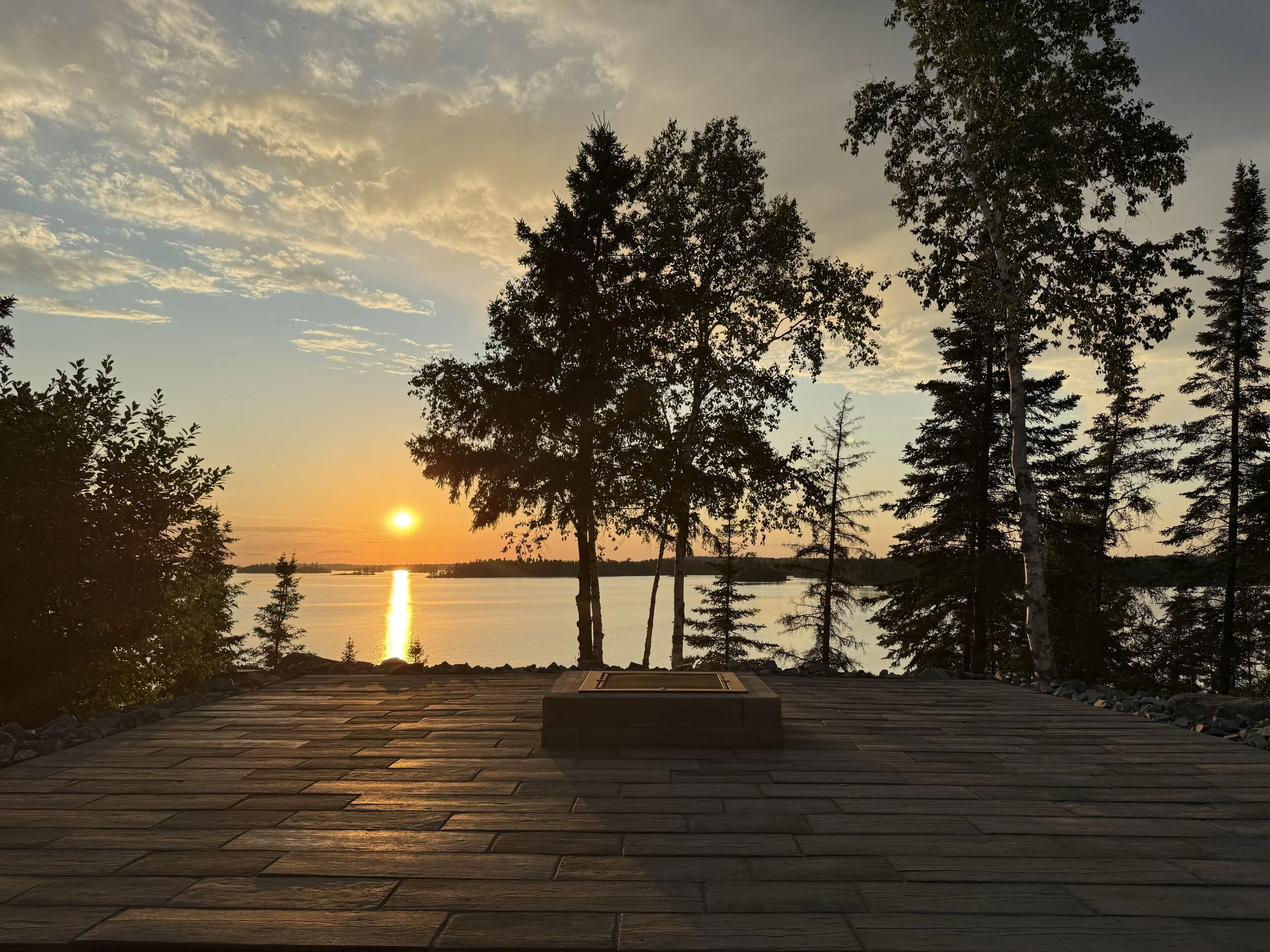 Sunset over a tranquil lake viewed from a wooden deck surrounded by trees.