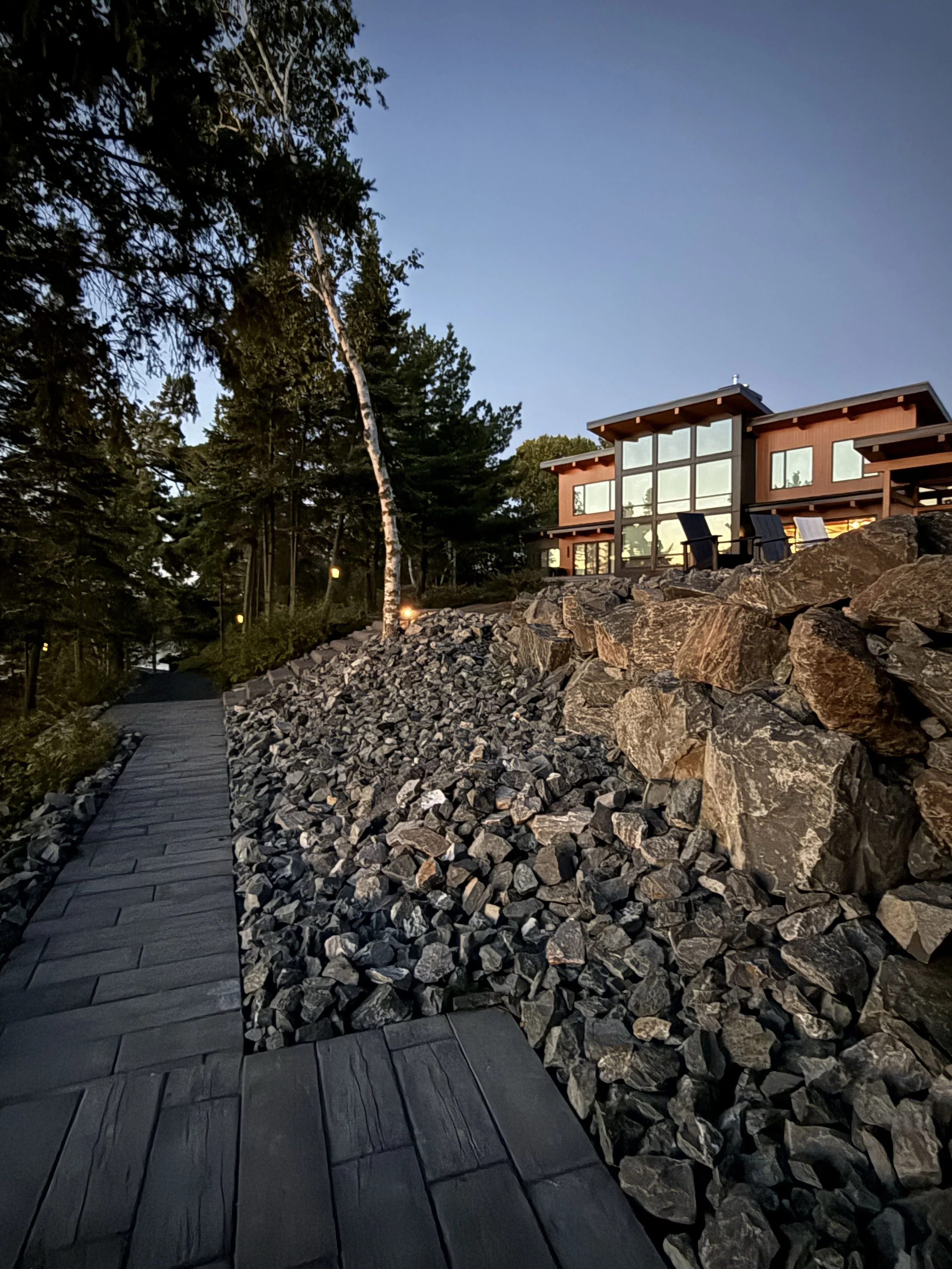 A modern house on a hill with large glass windows surrounded by rocks and trees, with a wooden pathway leading up to it during dusk.