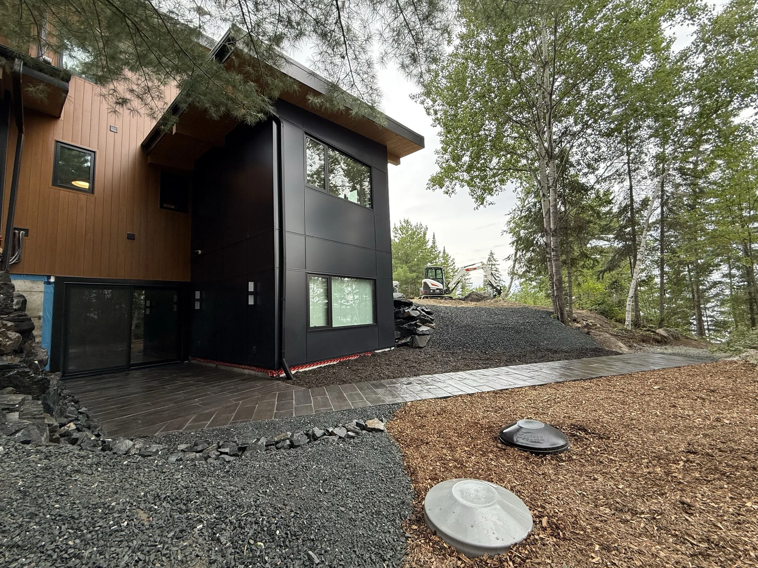 Modern house exterior with black and wood siding, large windows, and landscaped yard with trees and construction equipment in the background.