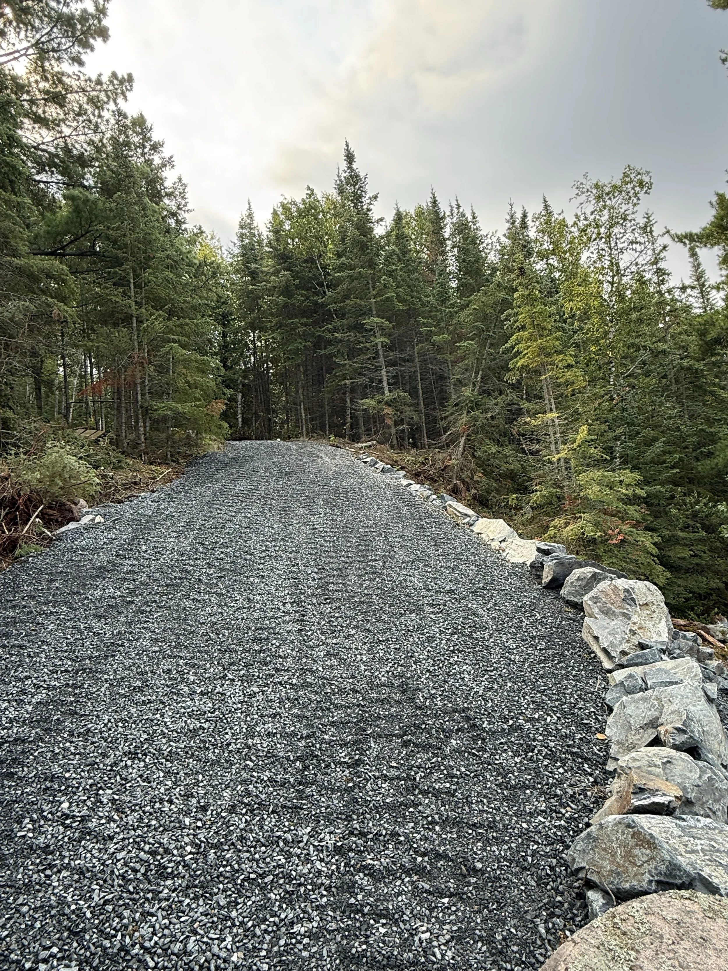 Gravel pathway lined with large rocks on the right side, surrounded by dense green forest under a partly cloudy sky.
