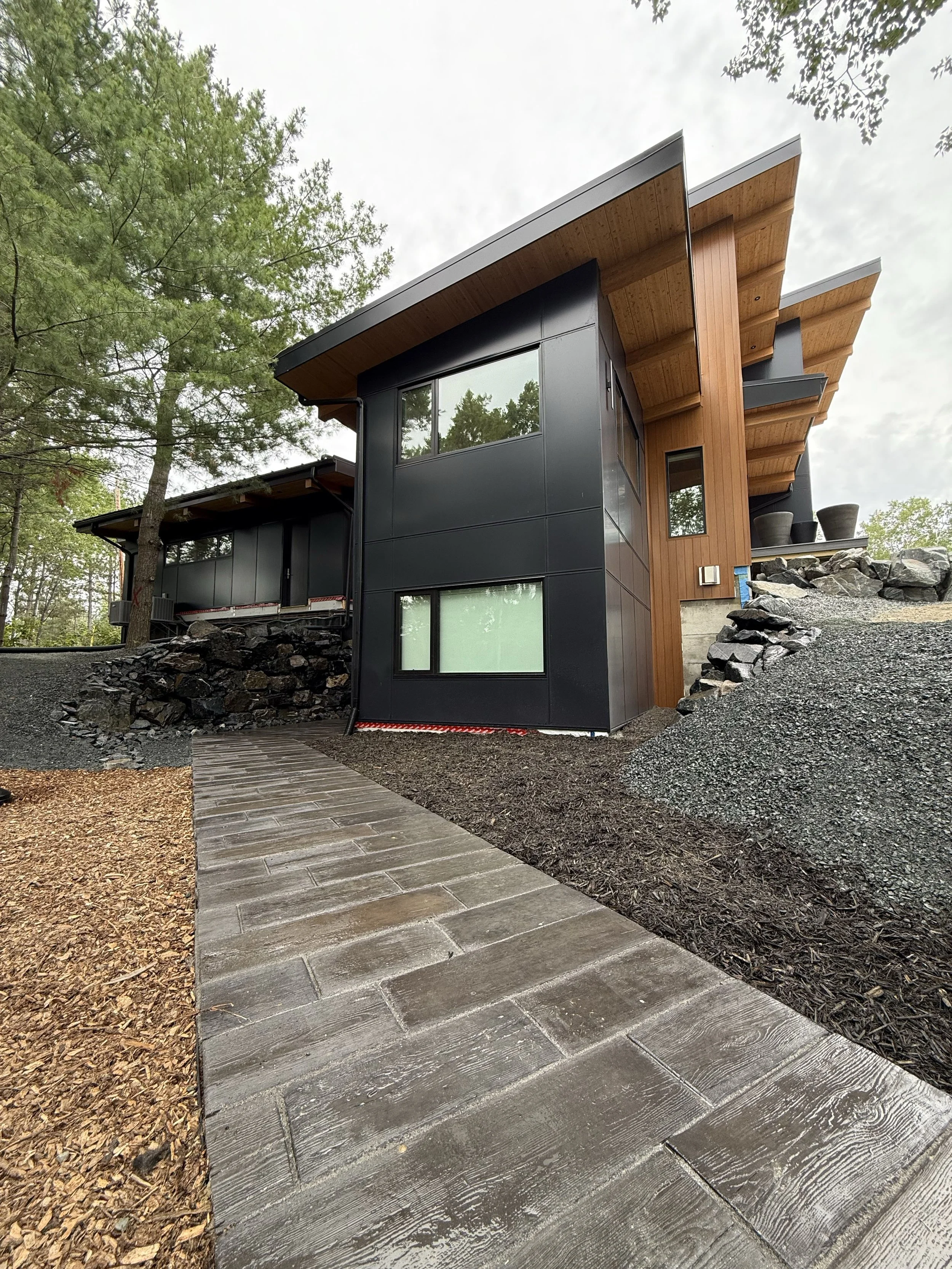 Modern house with black and wood exterior, large windows, surrounded by trees, gravel, and a stone pathway