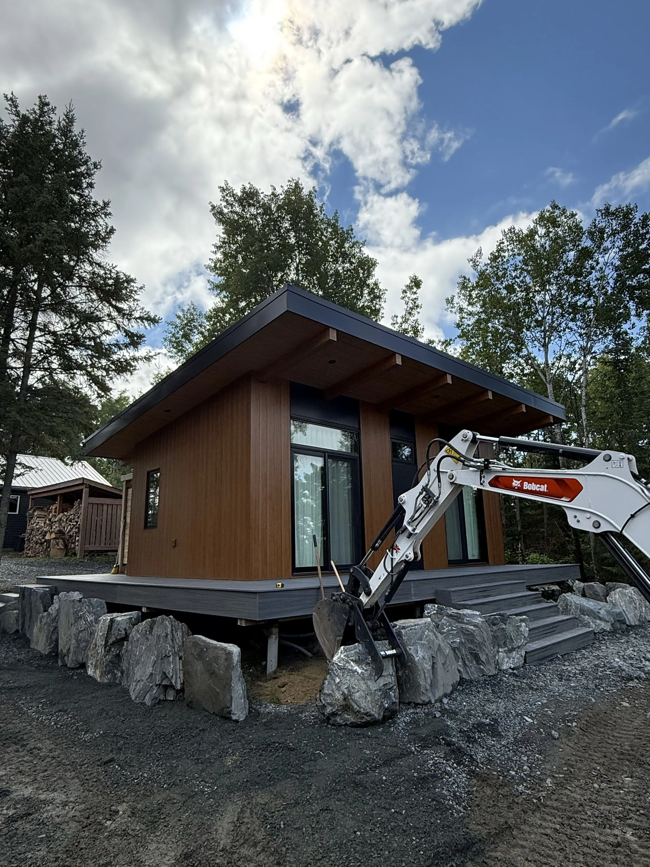 A modern small house with wooden exterior siding and large glass windows, built on a raised foundation with stones around its base, a small staircase leading to the entrance, and a construction excavator nearby, set against a background of a partly c