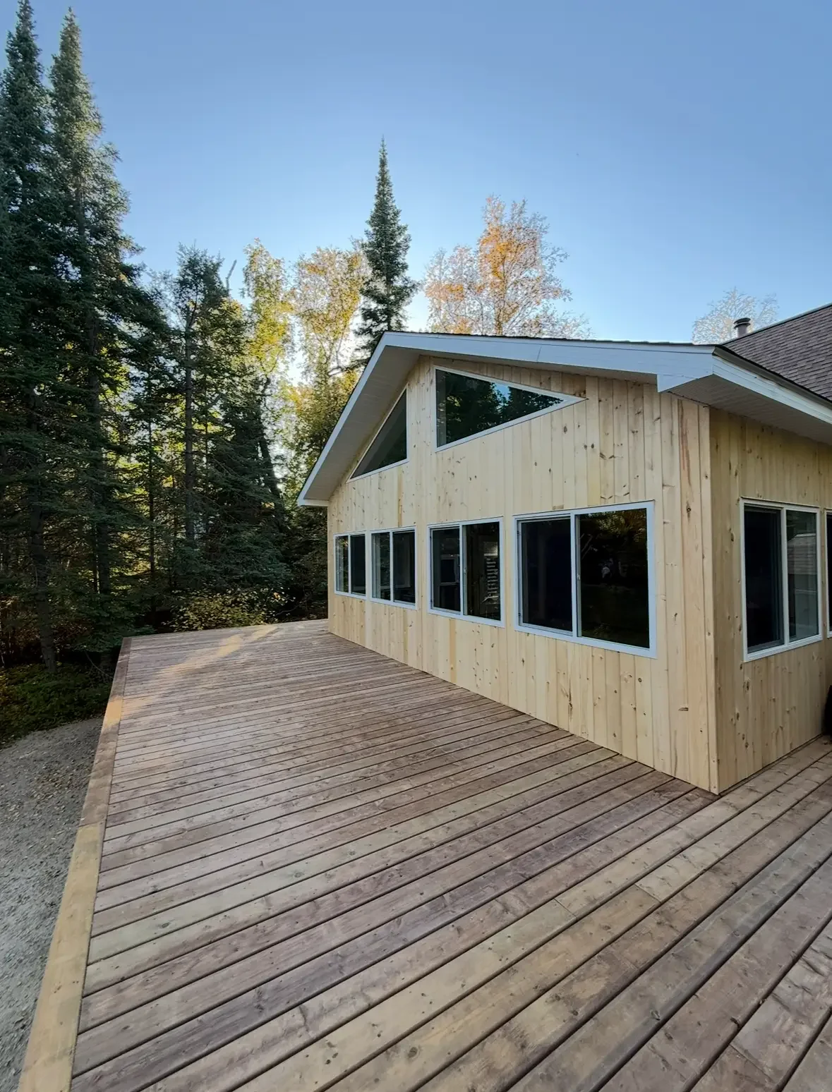 A modern house with a wooden exterior and multiple large windows, situated next to a forested area, featuring a spacious wooden deck.
