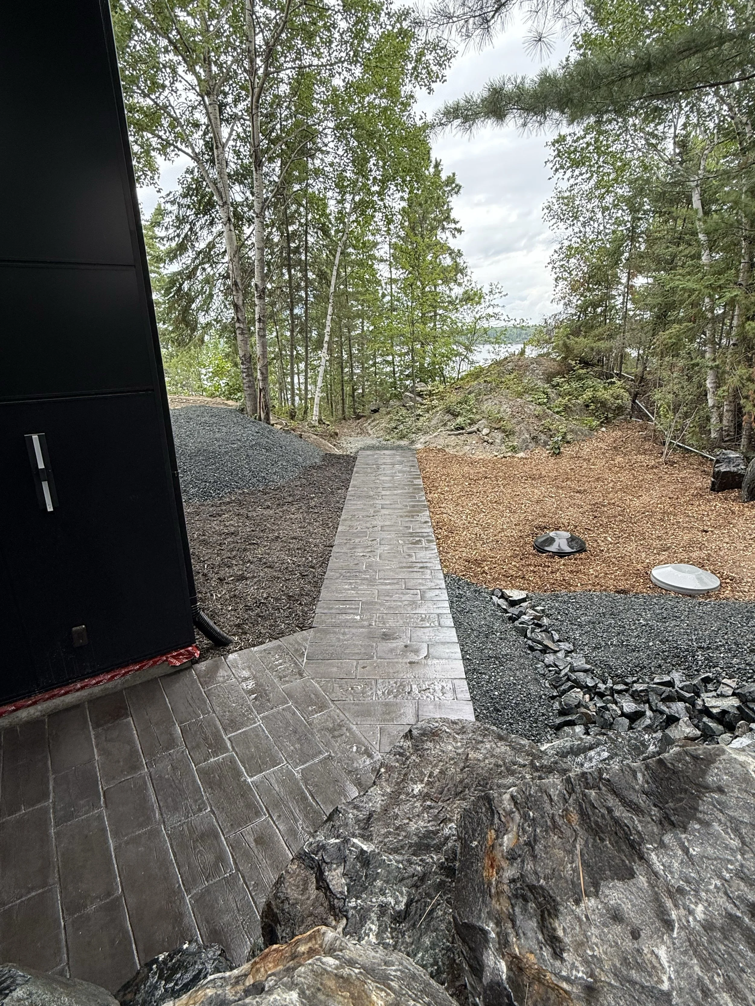 View of a landscaped backyard with a paved stone pathway leading towards a wooded area near a body of water, surrounded by trees and rocks.