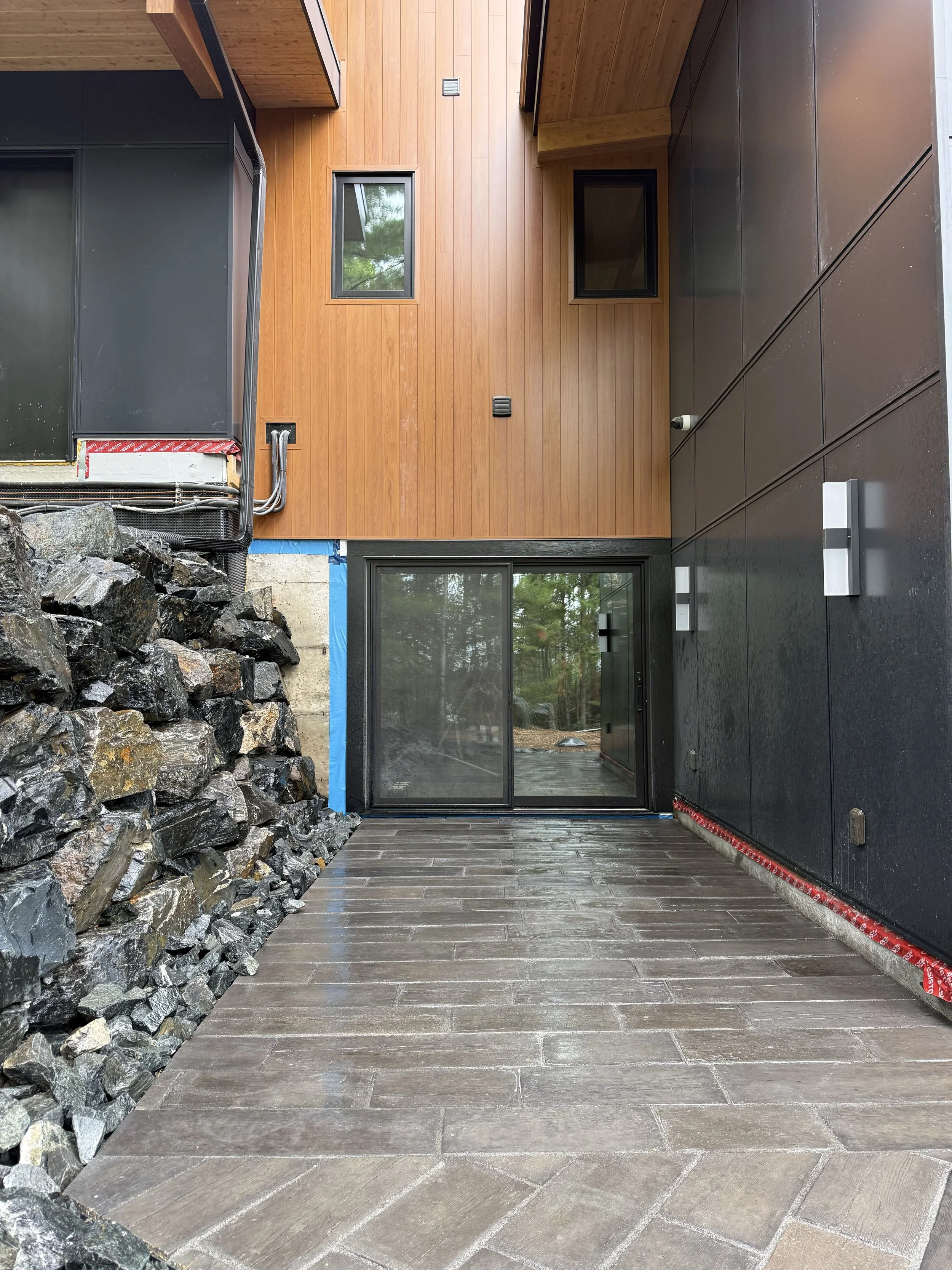 Modern house exterior with wooden and black siding, sliding glass door, stone accent wall, and paved patio.