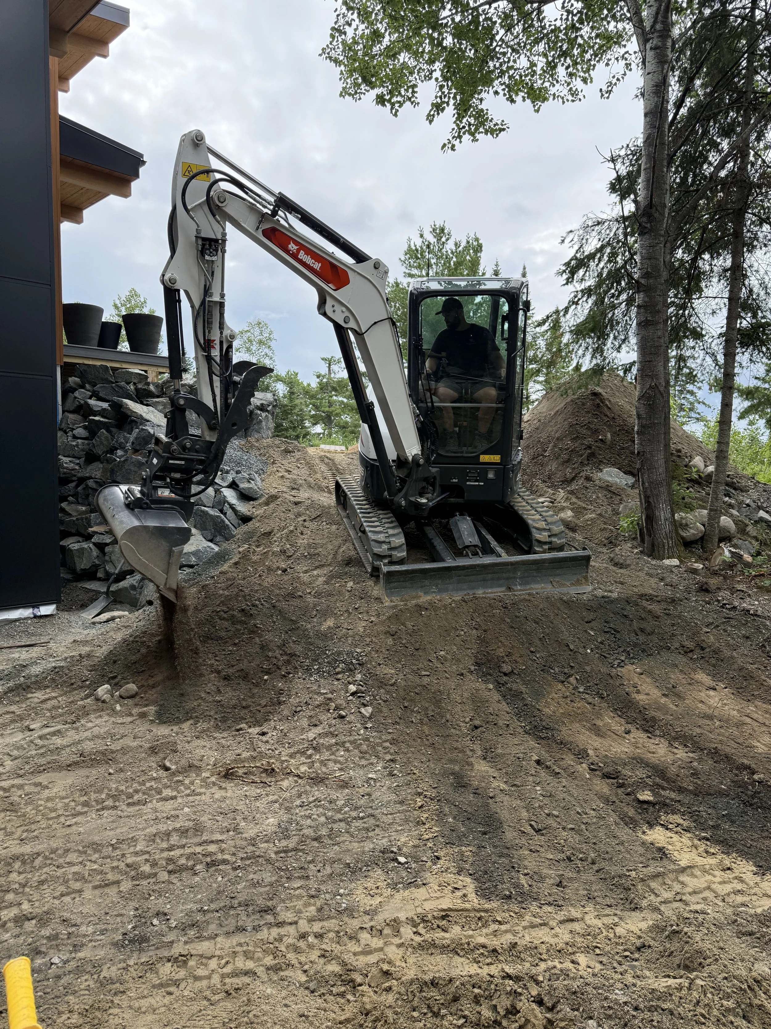Small Bobcat excavator moving dirt on a construction site, surrounded by trees and rocks.