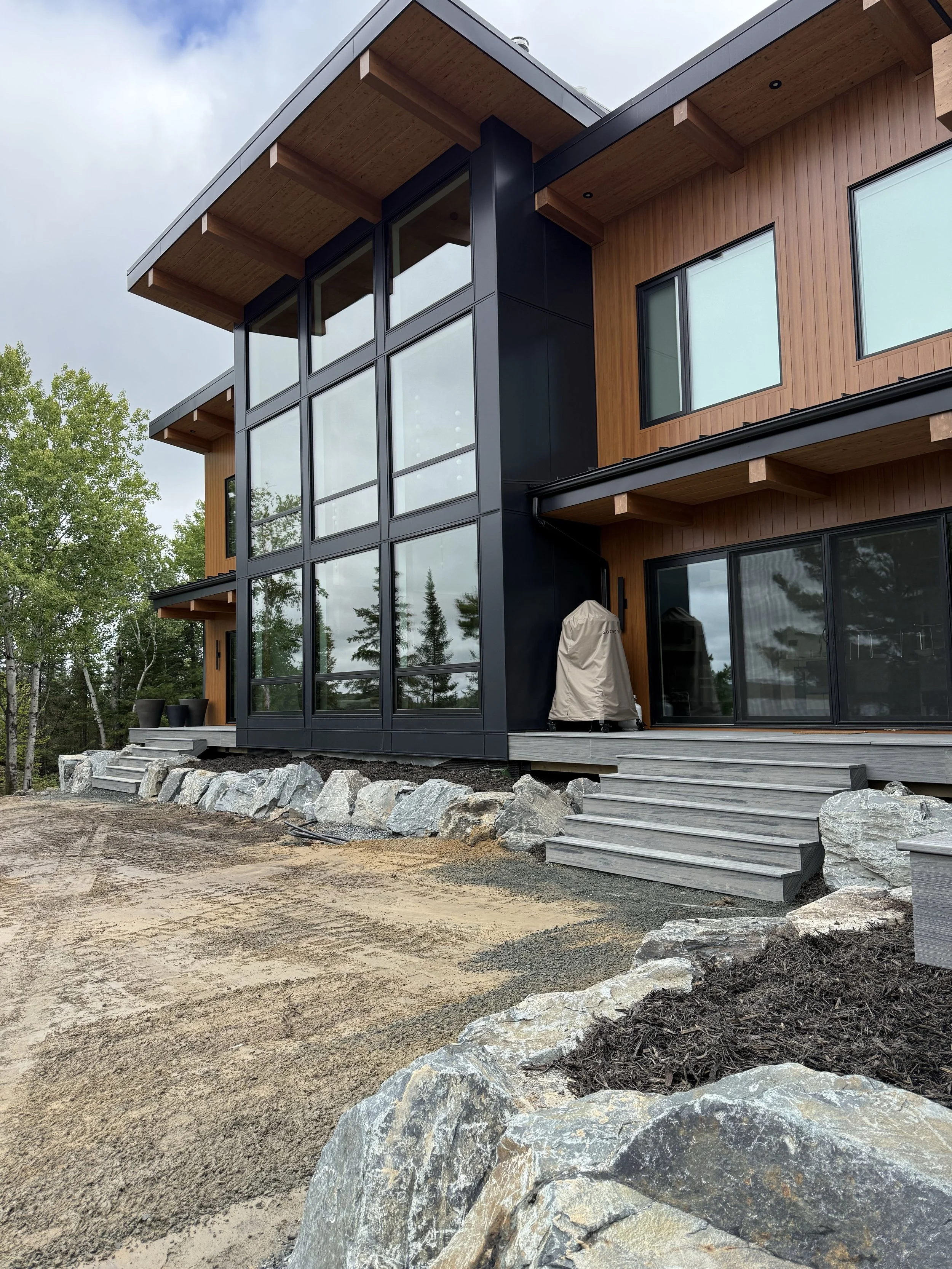 Modern multi-story house with large glass windows, wooden exterior, and stone landscaping, situated in a natural setting with trees.
