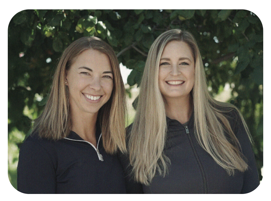 Two smiling women with long hair, wearing black zip-up sports jackets, standing outdoors in front of green leafy trees.