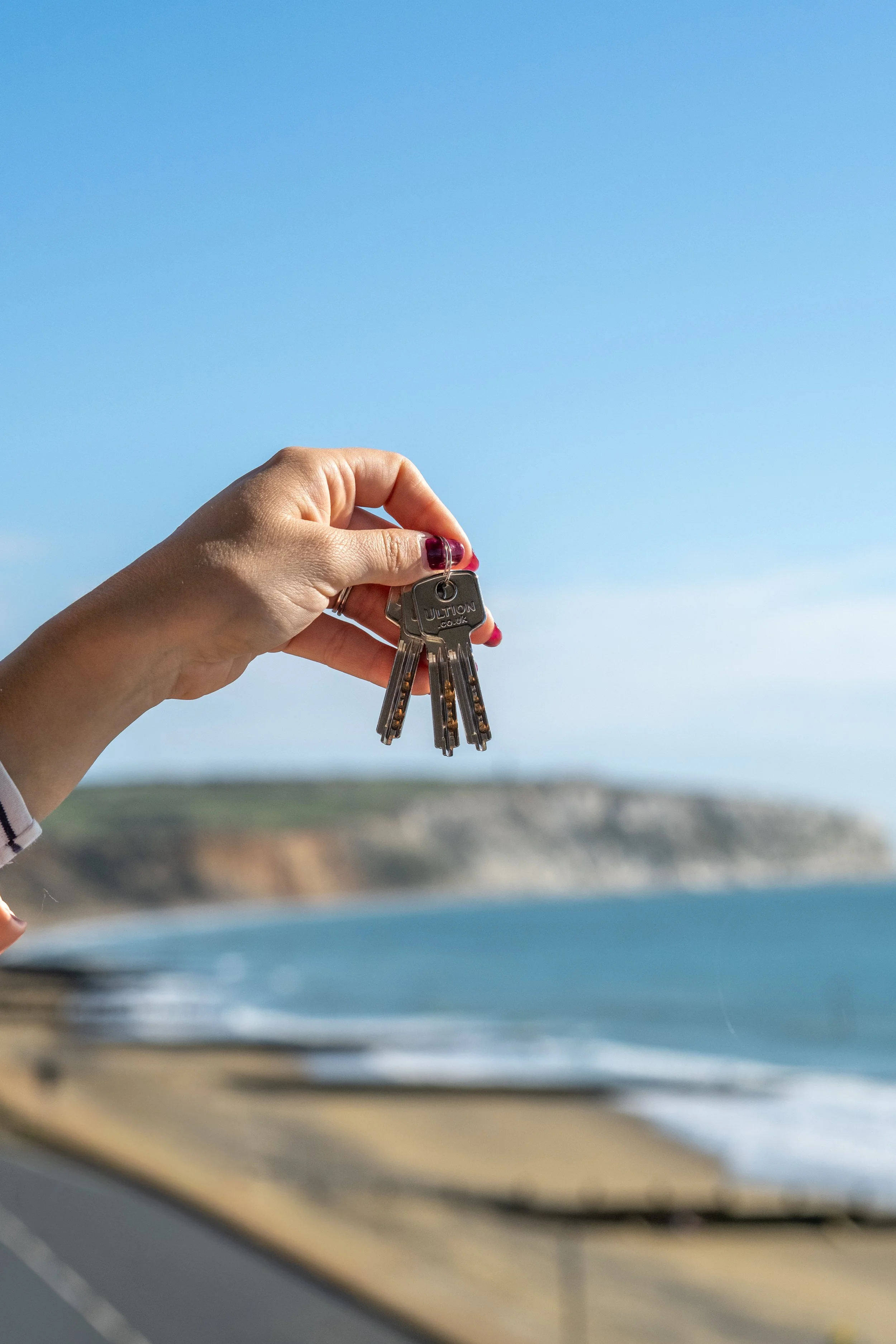 A hand holding a set of keys with a beach and coastline in the background.