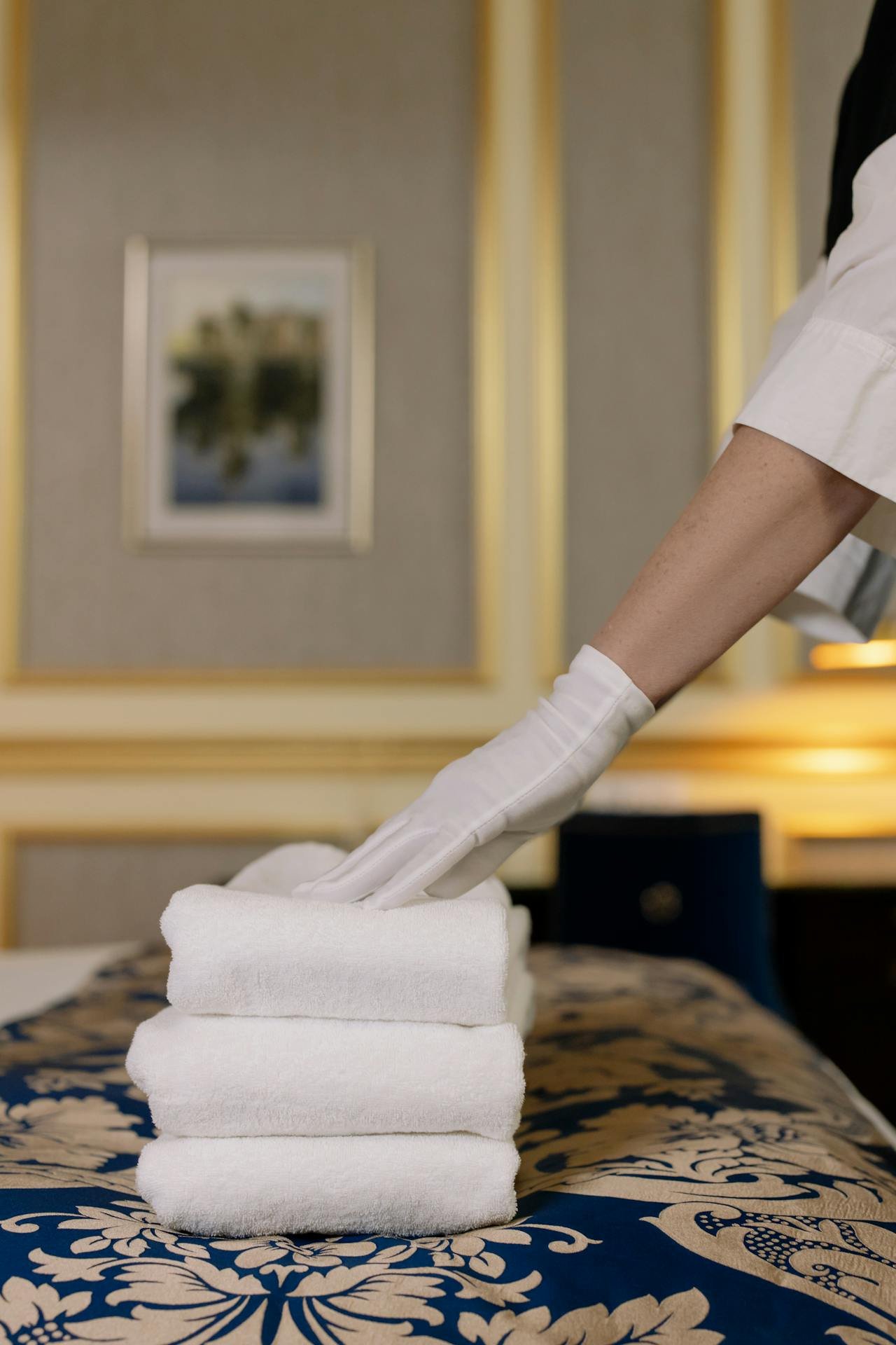 A stack of clean towels in a hotel guest room and a housekeepers hands wearing gloves smoothing them out.