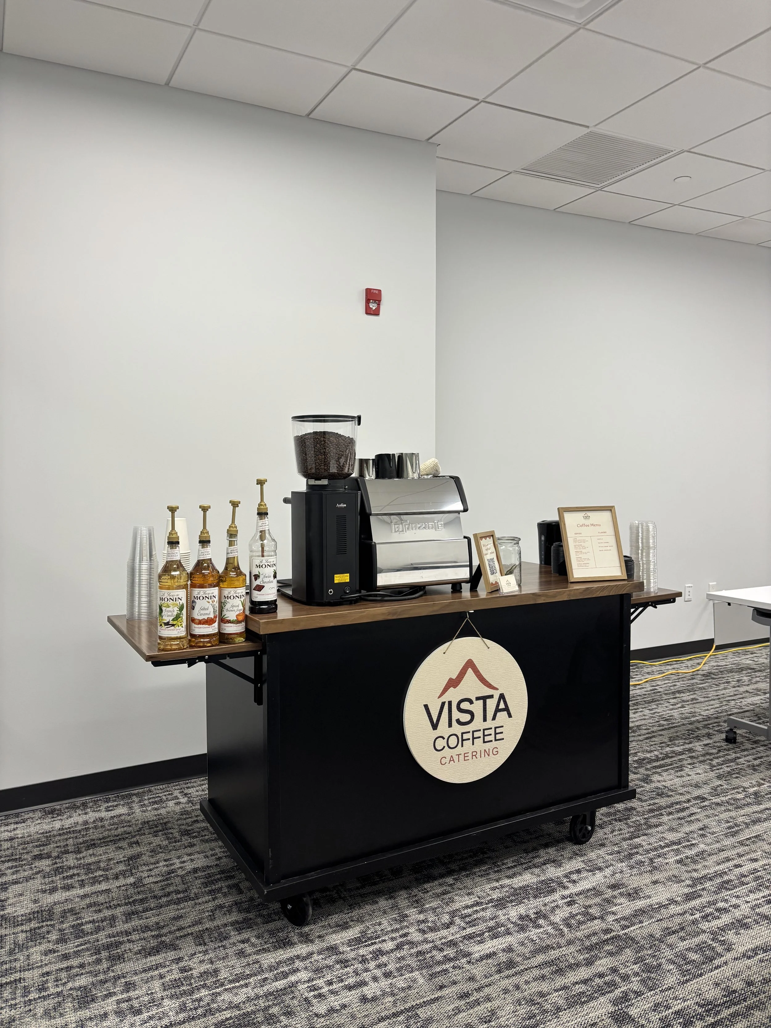 Coffee station with a coffee machine, bottles of Monin syrup, disposable cups, and a sign for Vista Coffee Catering in an office setting.