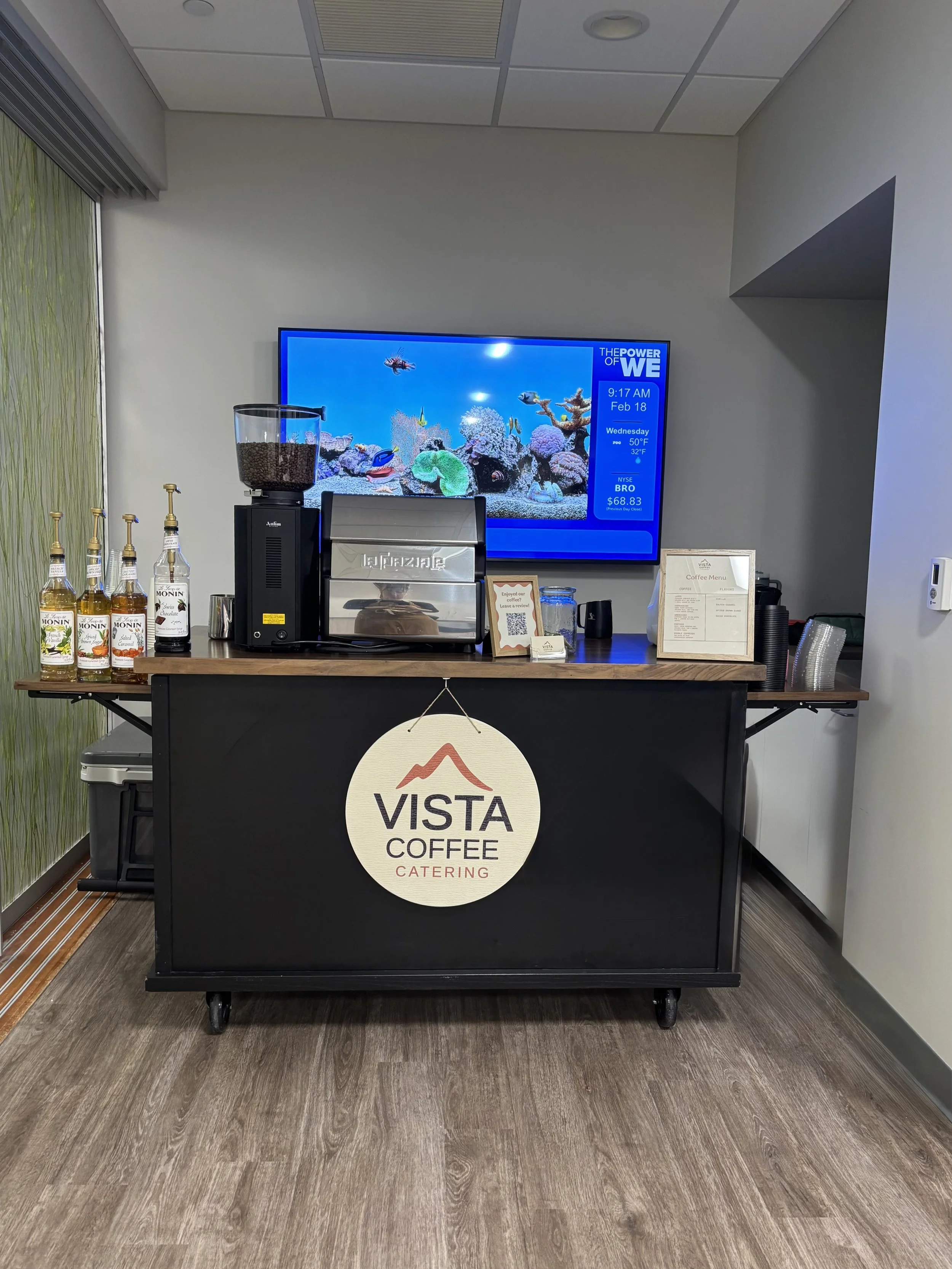 A coffee station on a black cart with a "Vista Coffee Catering" sign, a coffee grinder, a coffee machine, a jar of coffee beans, and bottles of Monin syrup. Behind the station is a wall-mounted digital display screen showing an aquarium with fish, th
