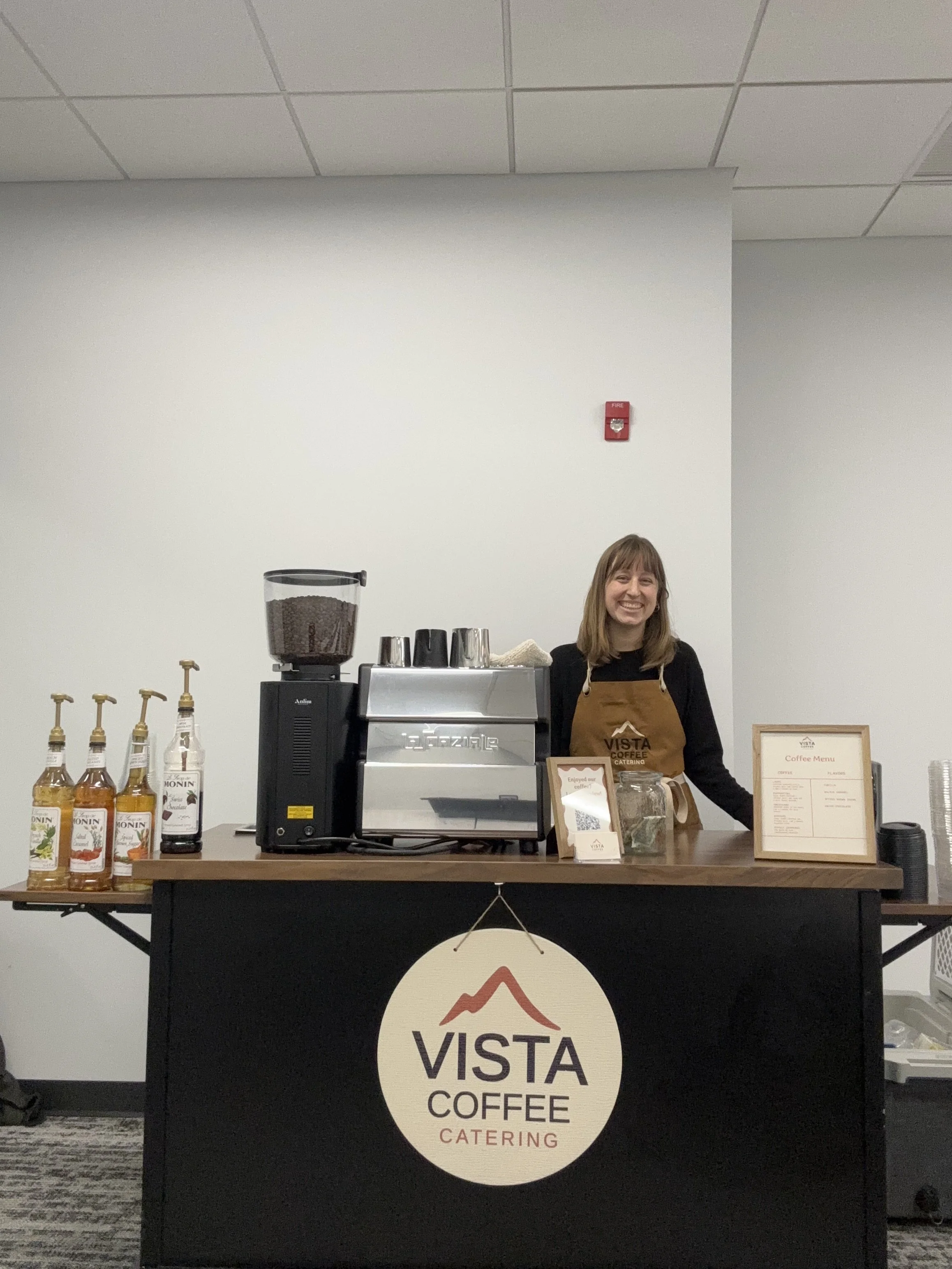 A smiling woman standing behind a coffee counter with a large coffee machine, bottles of syrup, and a menu board, at Vista Coffee Catering.