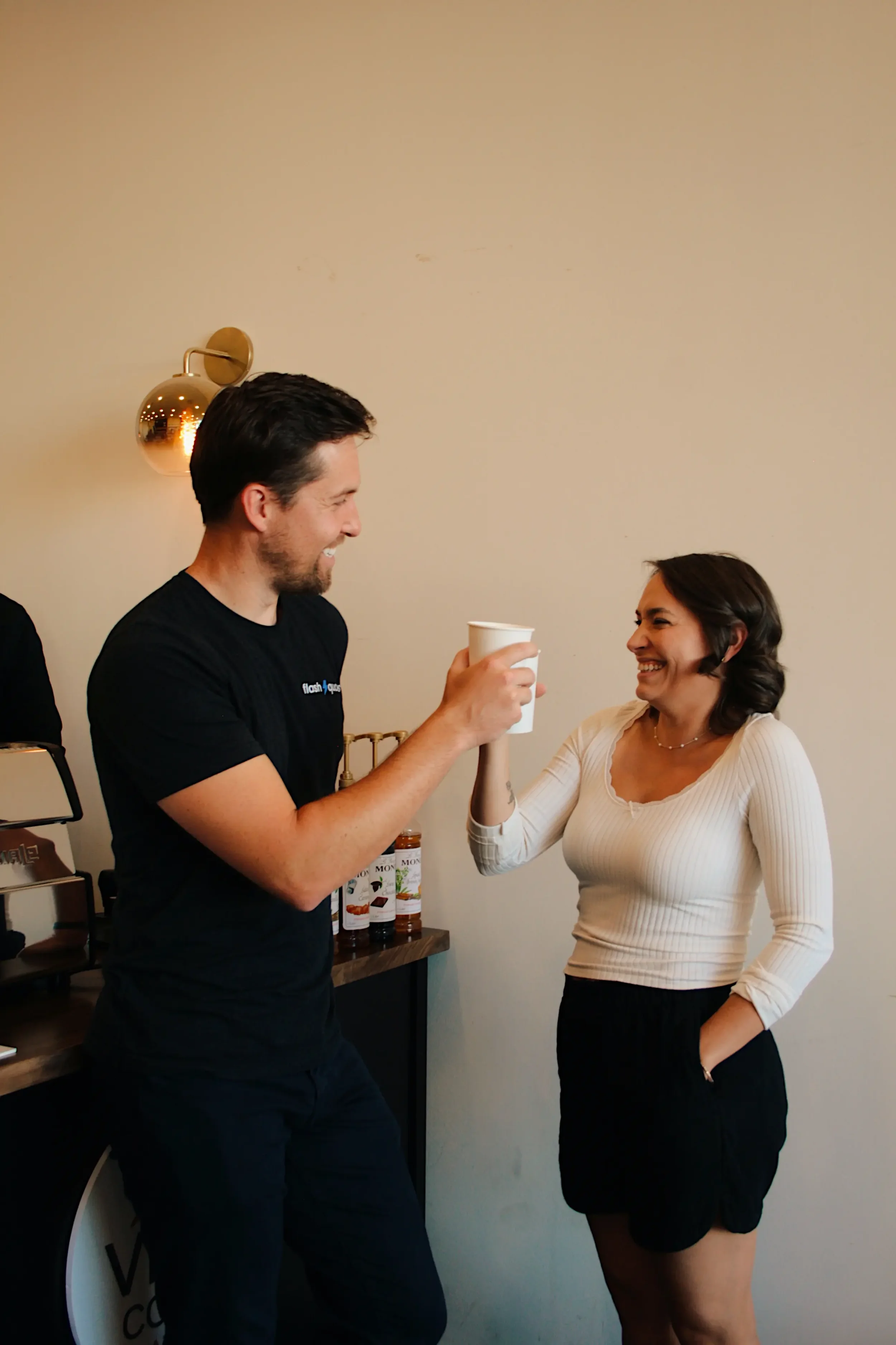 A man and woman smiling and clinking coffee cups inside a room with a lamp and bottles in the background.
