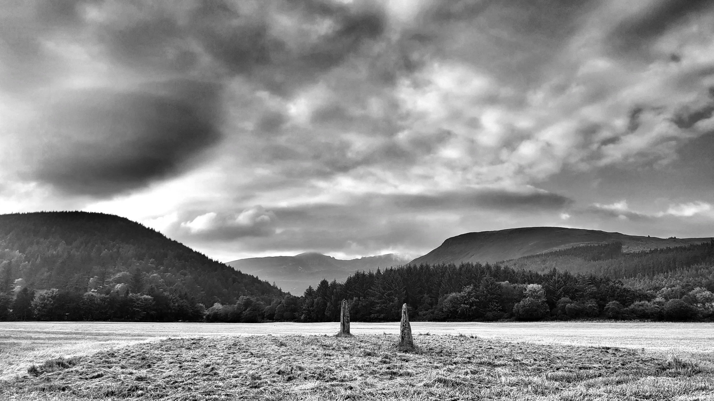Black and white landscape photo of a wide grassy field, two standing stones in the center, dense trees in the background, hills, and mountains under a cloudy sky.