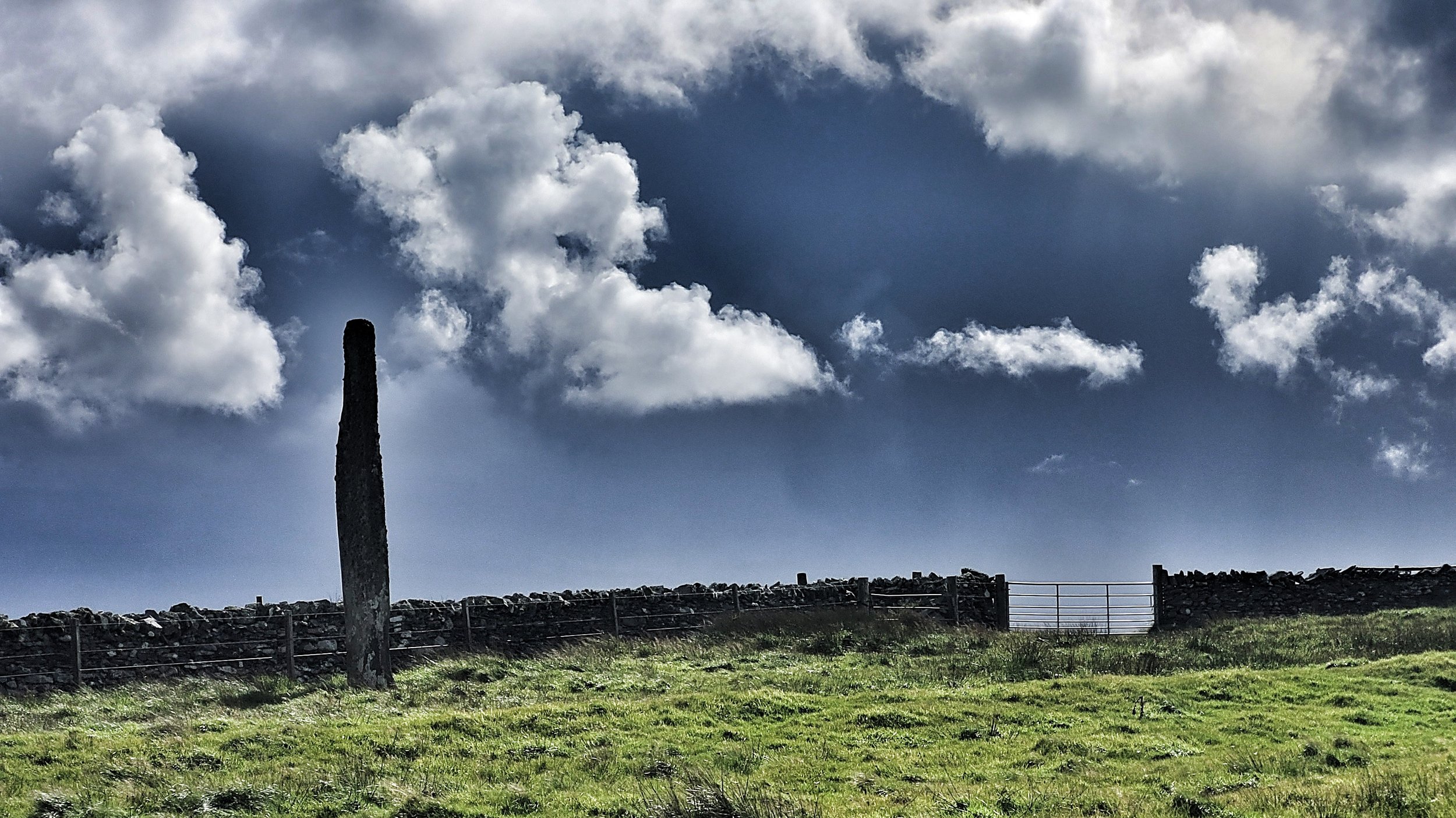A grassy field with a stone fence and a wooden post against a cloudy sky.