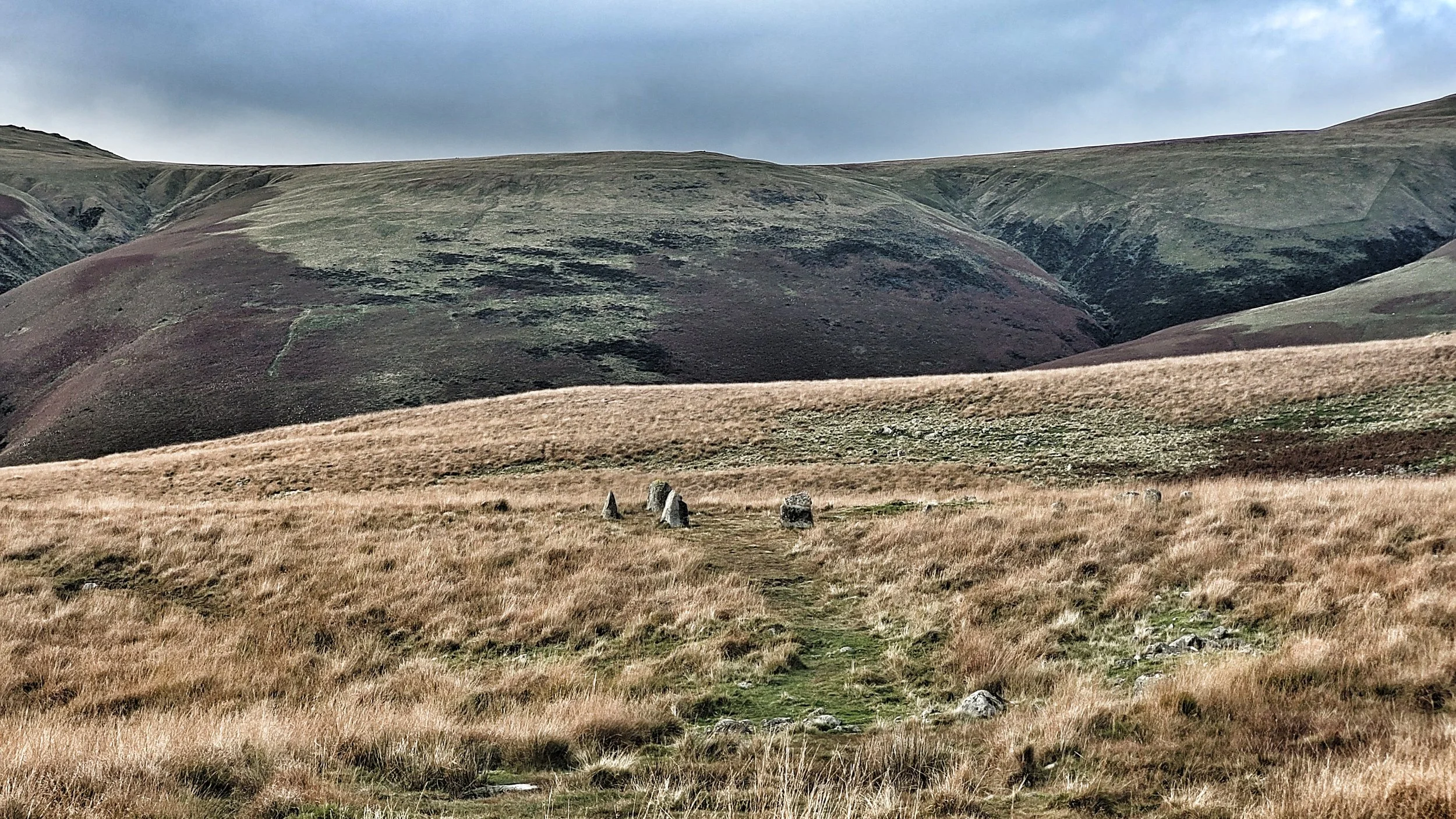 A wide open grassy hillside with three standing stones forming a small circle, and large rolling hills in the background under a cloudy sky.