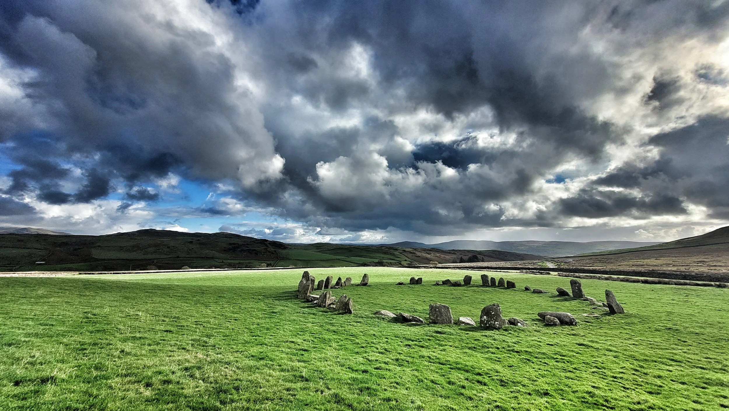 Green grassy field in front of a historic stone circle, with rolling hills and a cloudy sky overhead.