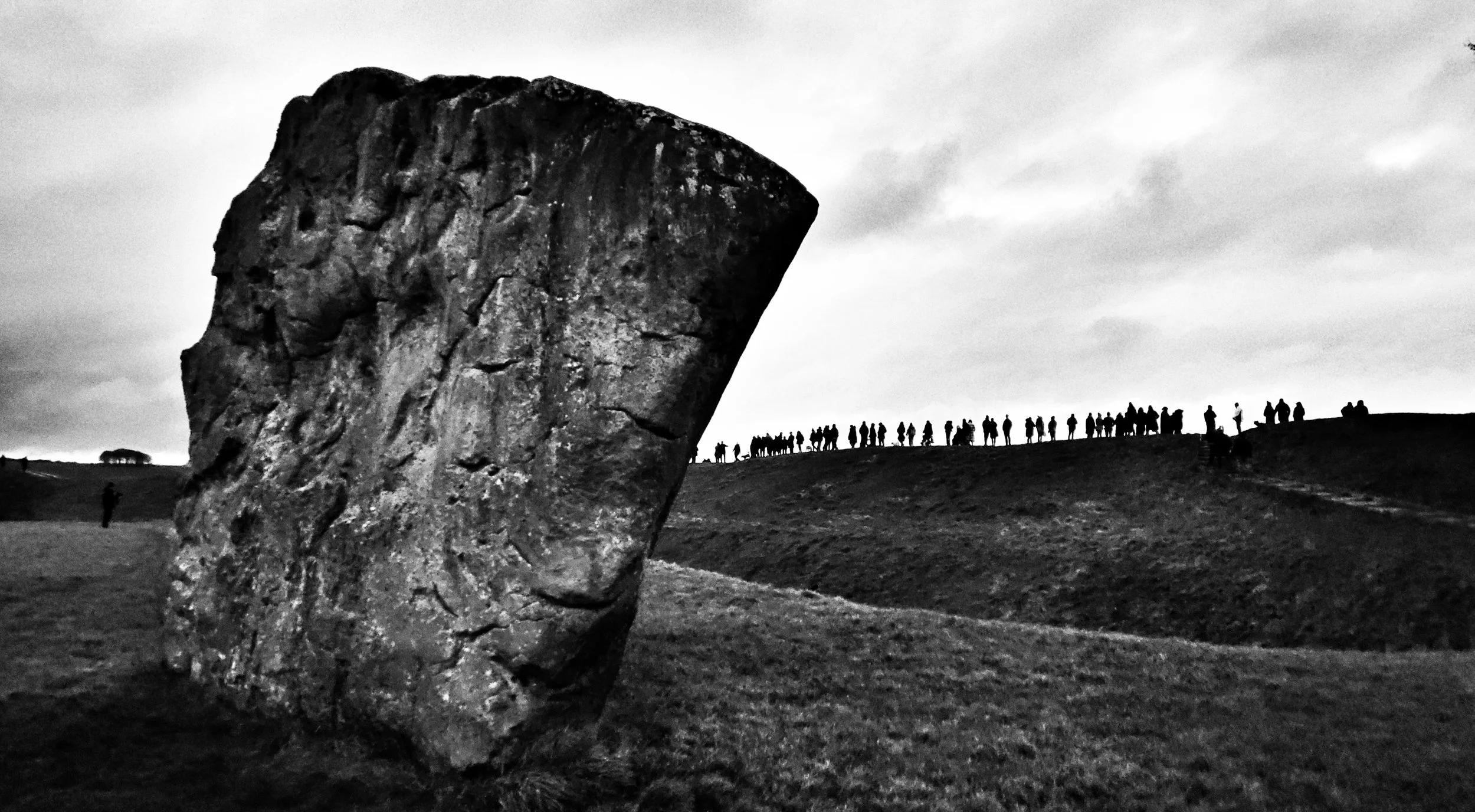 A large, weathered standing stone on a grassy landscape with people and horses in the background on a cloudy day.