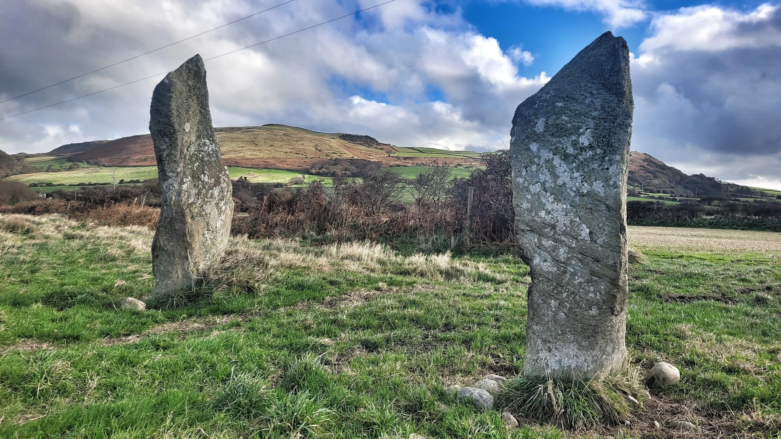 Two ancient standing stones in a grassy field with rolling hills and cloudy sky in the background.