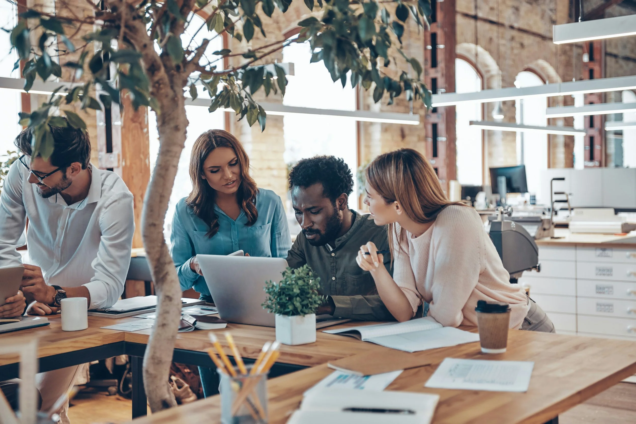 Group of four colleagues working together at a wooden table in a modern office, reviewing documents and using laptops. The office has exposed brick walls, plants, trees, large windows, and modern lighting.