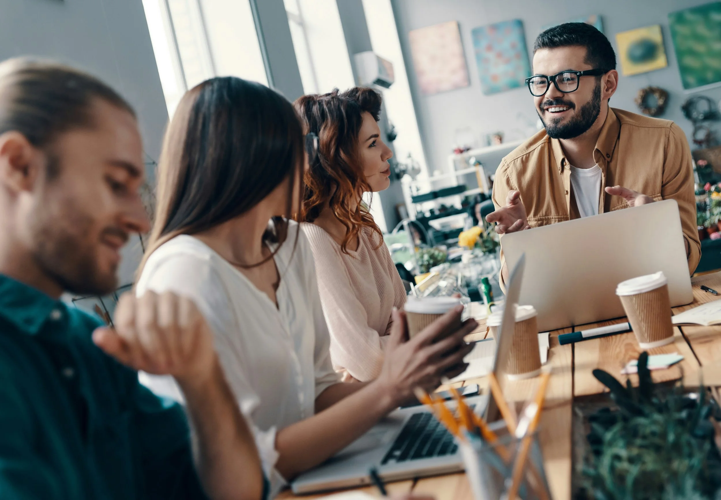 Group of four young adults chatting during a meeting in a bright office space with colorful artwork on the wall.