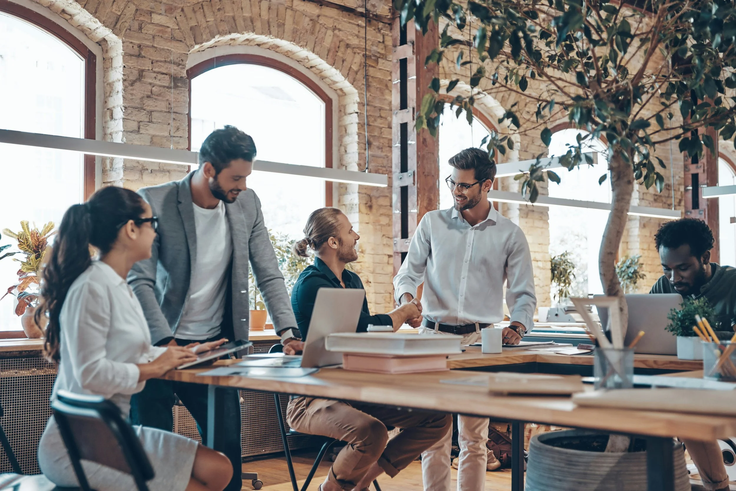 A group of five diverse professionals in a modern, well-lit office space, with three men and two women, engaged in a business meeting, smiling, and shaking hands.
