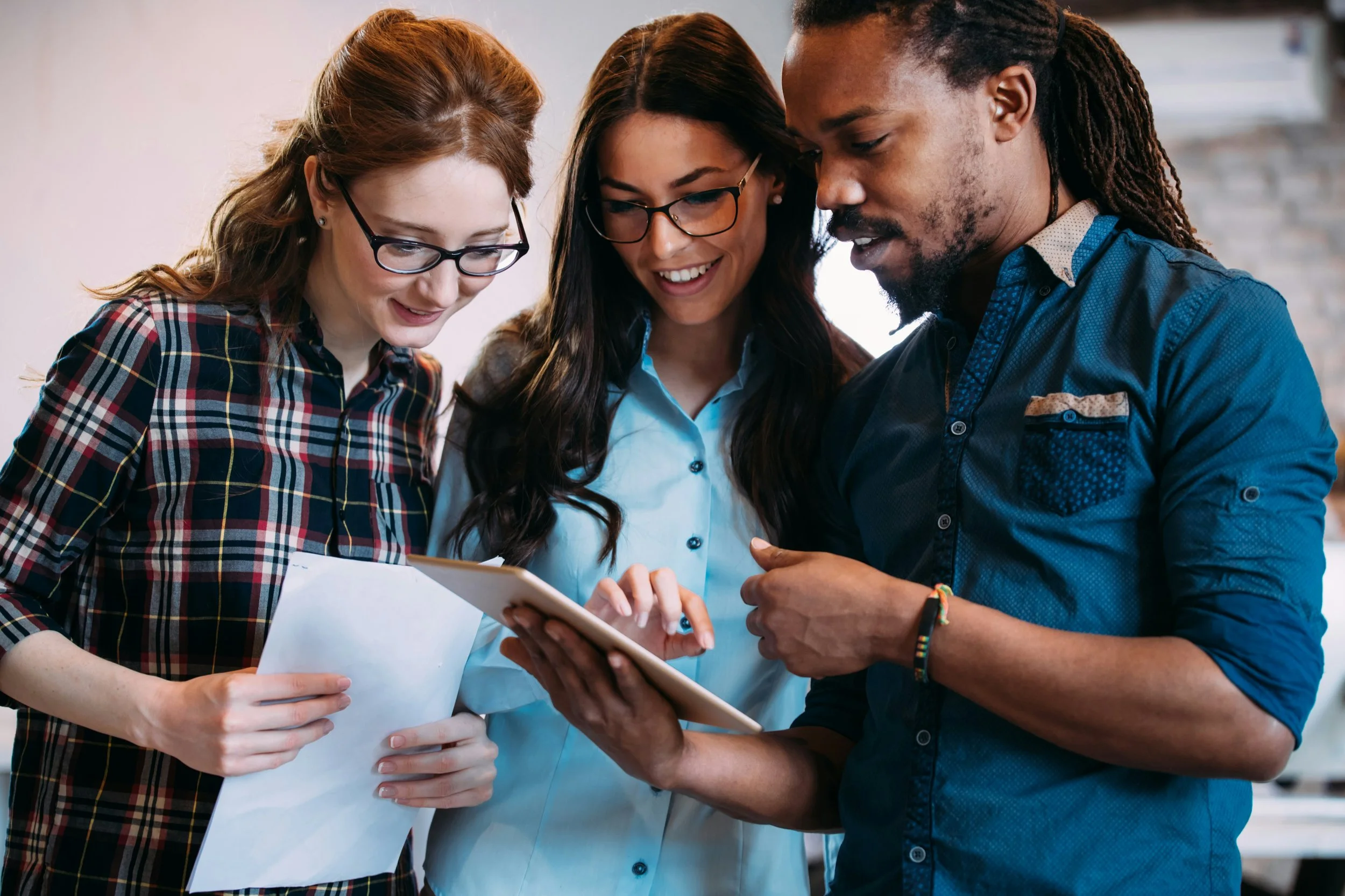 Group of four colleagues working together at a wooden table in a modern office, reviewing documents and using laptops. The office has exposed brick walls, plants, trees, large windows, and modern lighting.