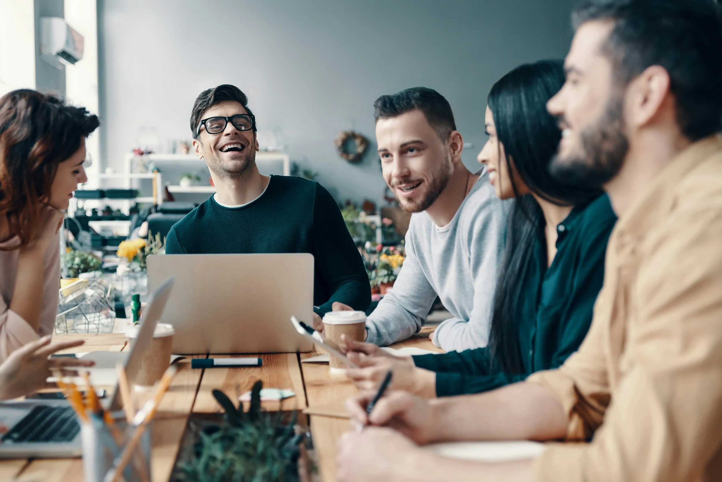 Five young adults sitting at a wooden table, engaging in a lively discussion with smiles in a bright, modern workspace or cafe with plants and decorations in the background.
