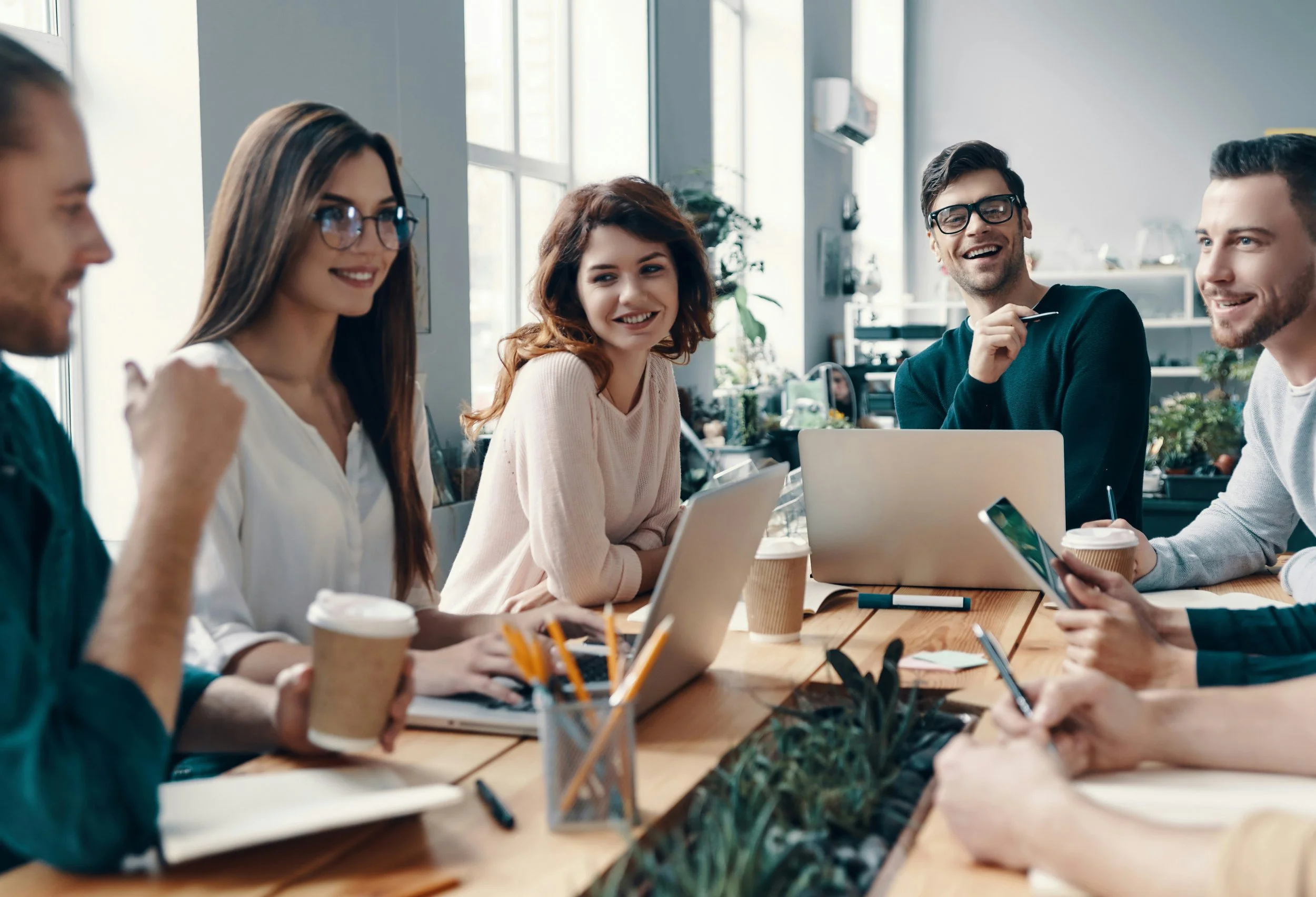 Group of five modern professionals sitting around a table in a bright contemporary office, engaged in a business meeting, with laptops and coffee cups.