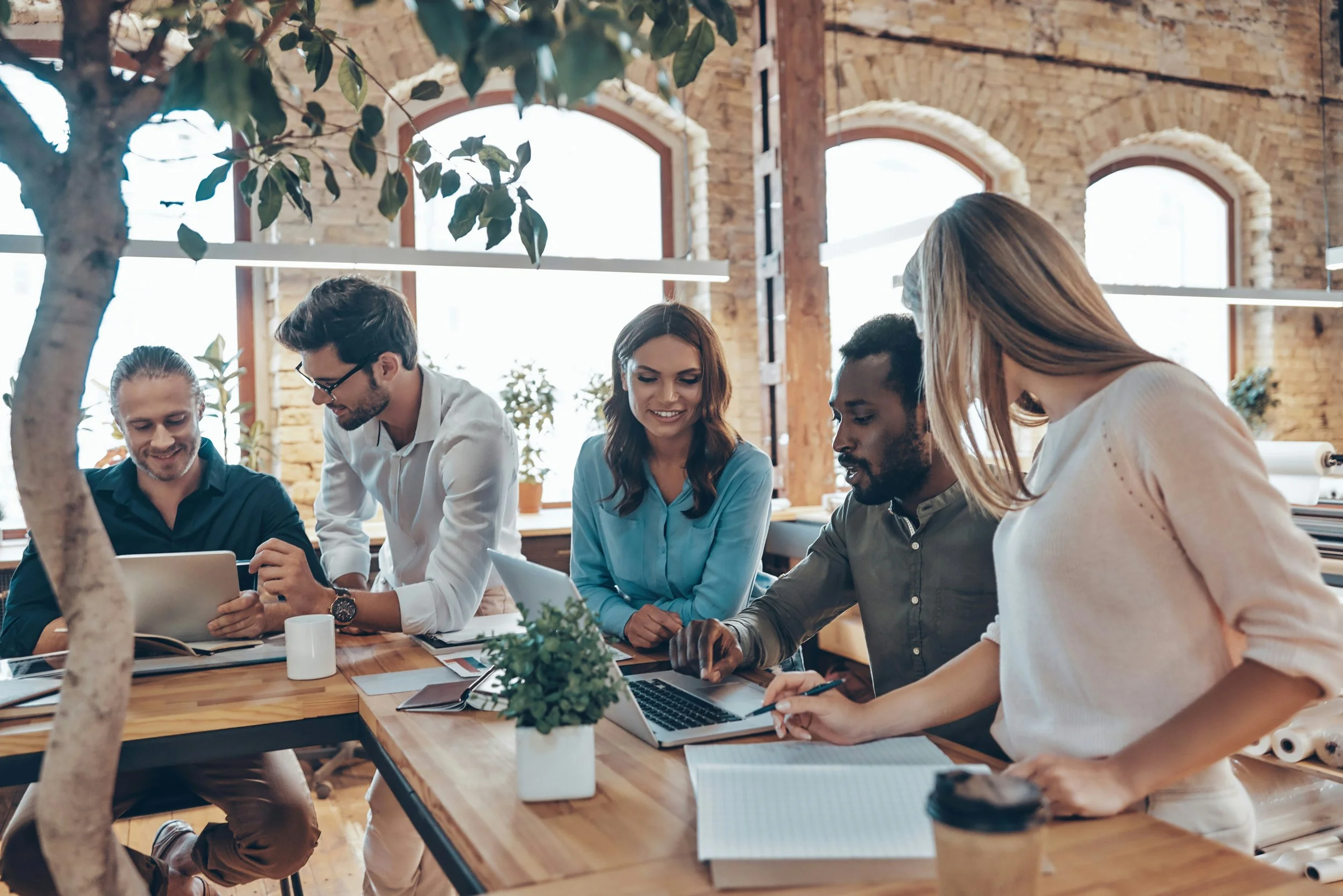Group of five diverse young professionals working together at a wooden table in a modern, brick-walled office with large windows and indoor plants.