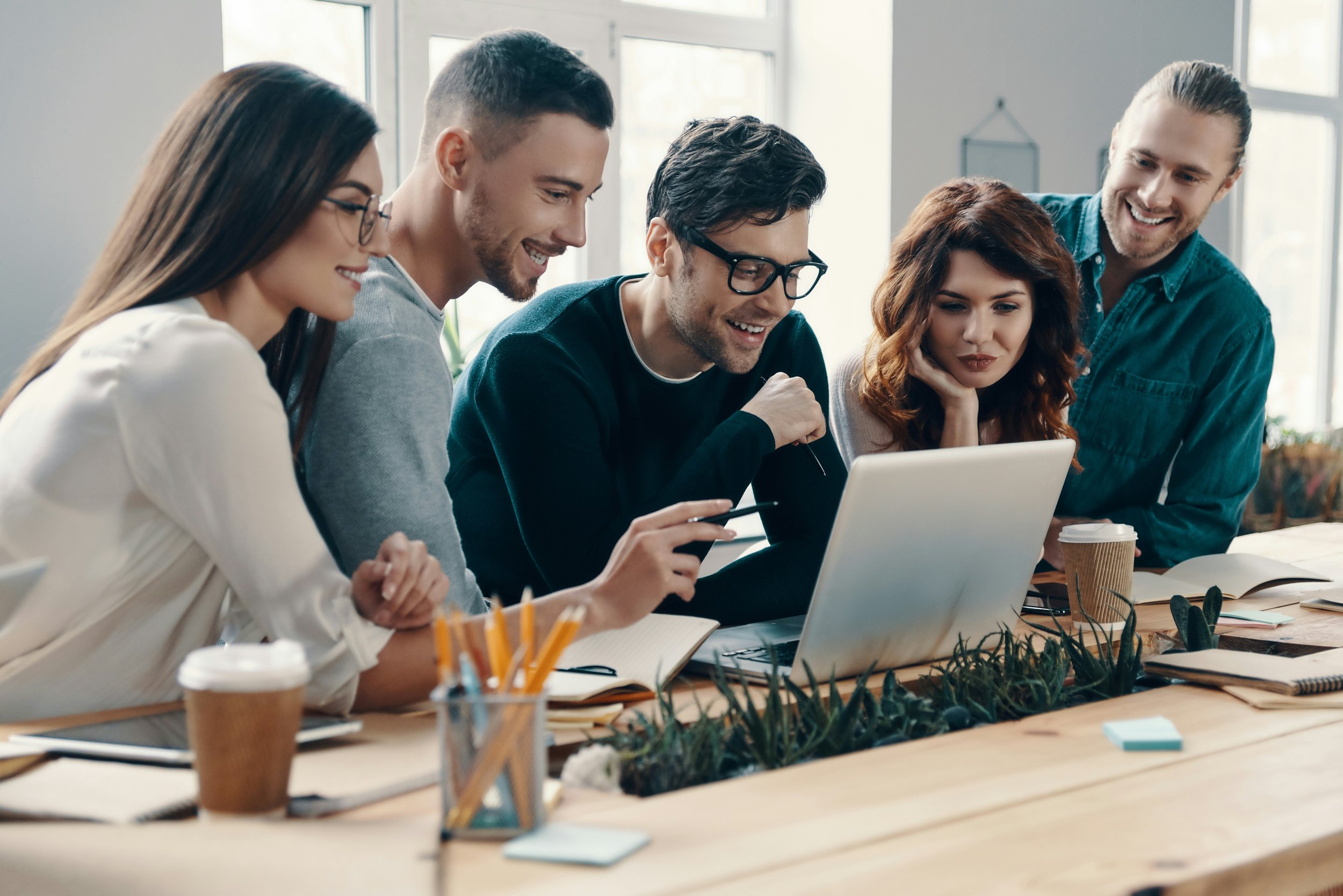 Five creative professionals gathered around a table looking at a laptop screen, smiling and engaging in a discussion, with notebooks, coffee cups, and office supplies on the table.