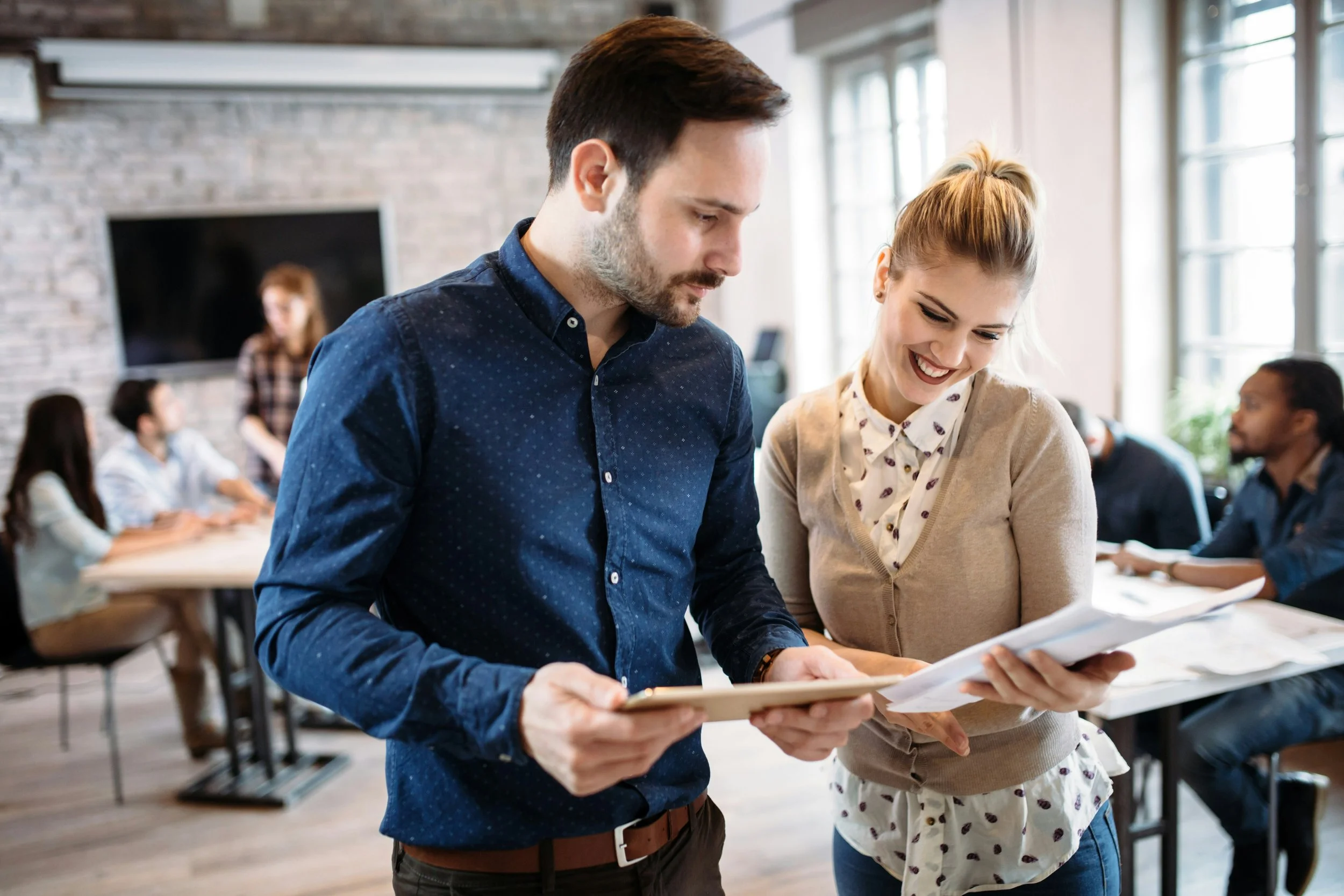 Six diverse people collaborating in a modern office, working on laptops and tablets, with notebooks and a plant on the table and a tree in the background.