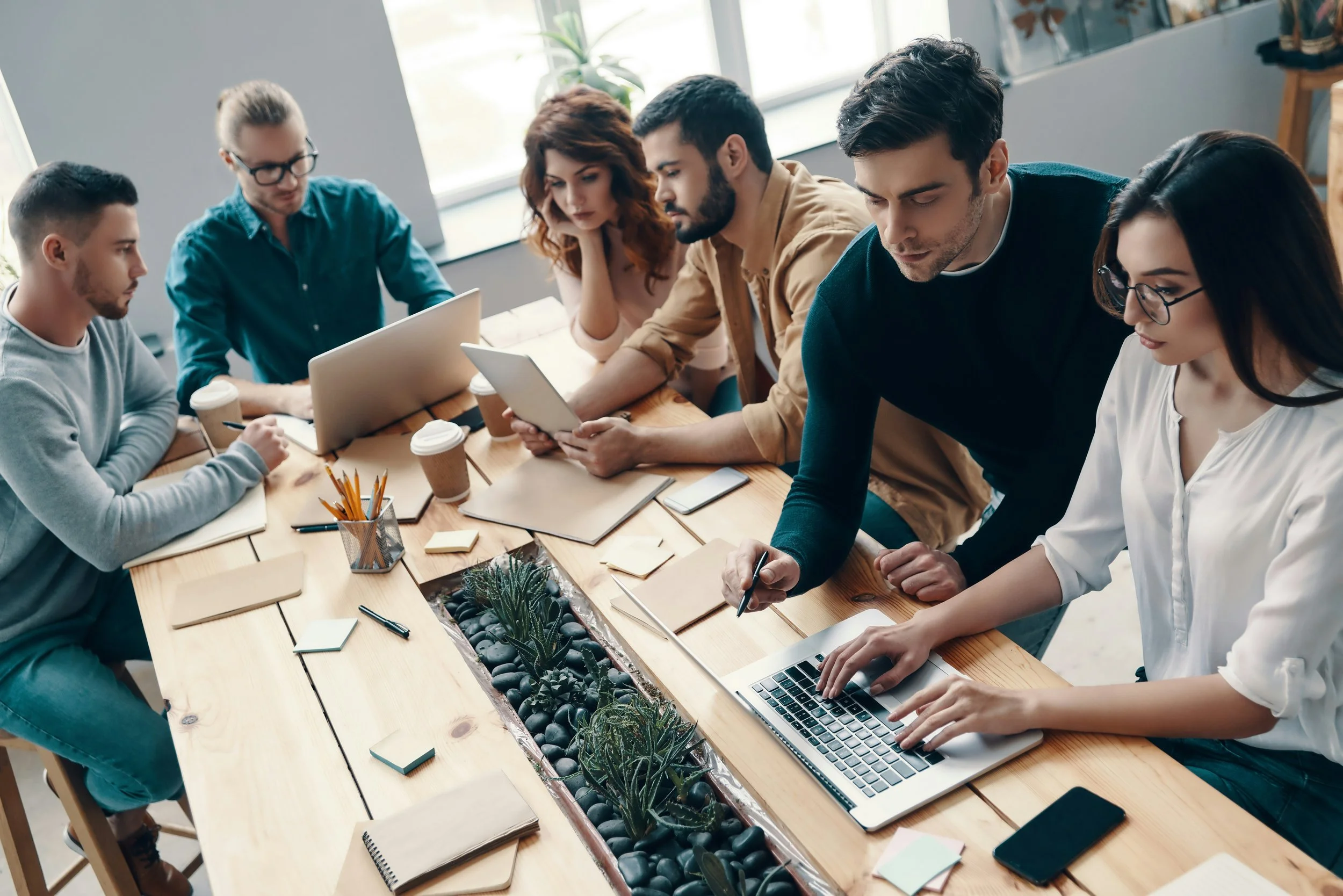 A group of six successful adults collaborating around a wooden table in a contemporary office space, some working on laptops and others using tablets or taking notes, with notebooks, coffee cups, and stationery on the table.