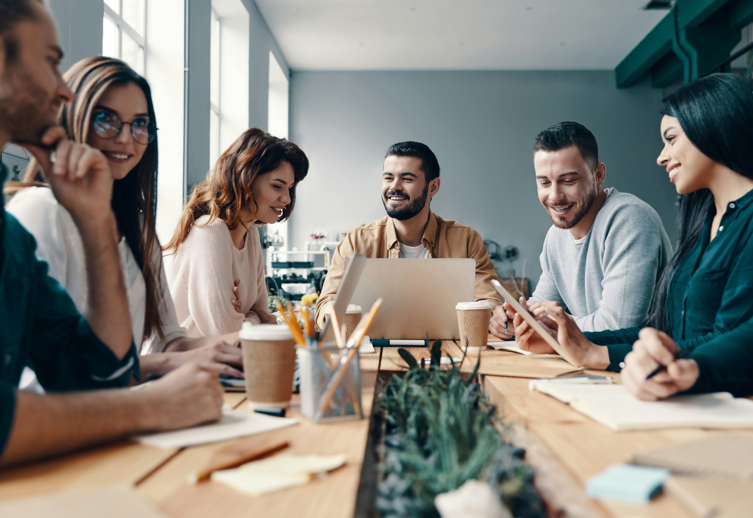 A diverse group of six professional people sitting around a conference table in a bright modern workspace, smiling and engaging in a meeting, with laptops, notebooks, and coffee cups on the table.