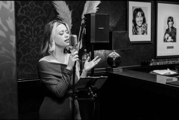 A woman singing into a vintage microphone in a music studio, with framed photos on the wall behind her.