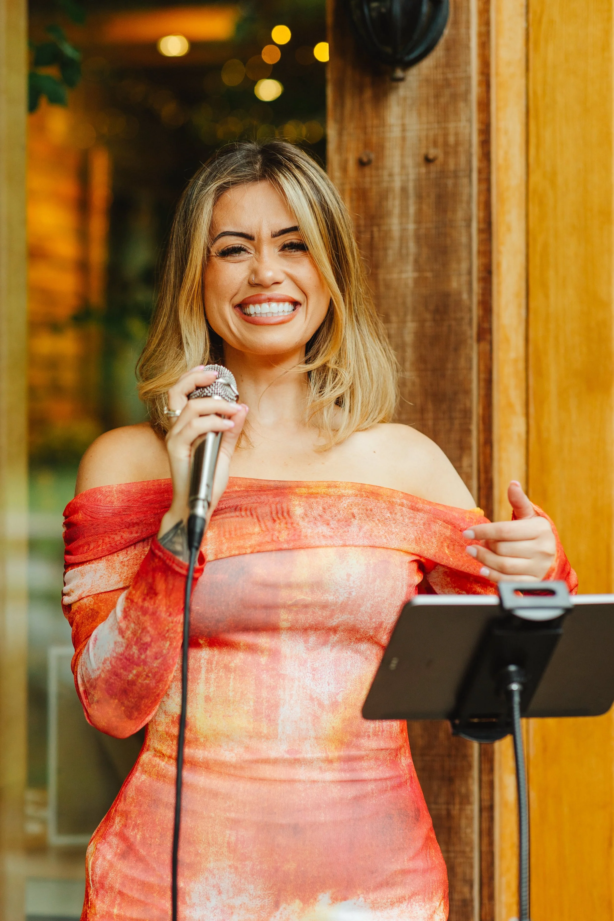 A woman smiling as she performs karaoke, holding a microphone with a music stand and tablet in front of her, with warm lighting and wooden wall background.