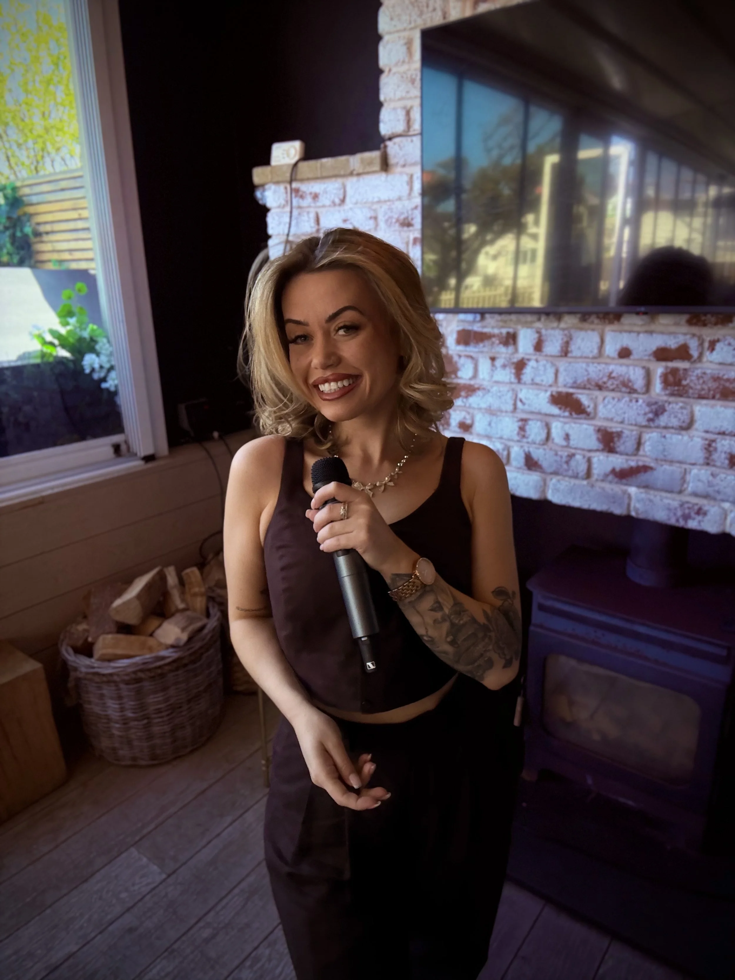 Woman with blonde hair holding a microphone, wearing a black sleeveless top and jewelry, standing indoors with a brick wall, a mounted TV, and a basket of wood logs in the background.
