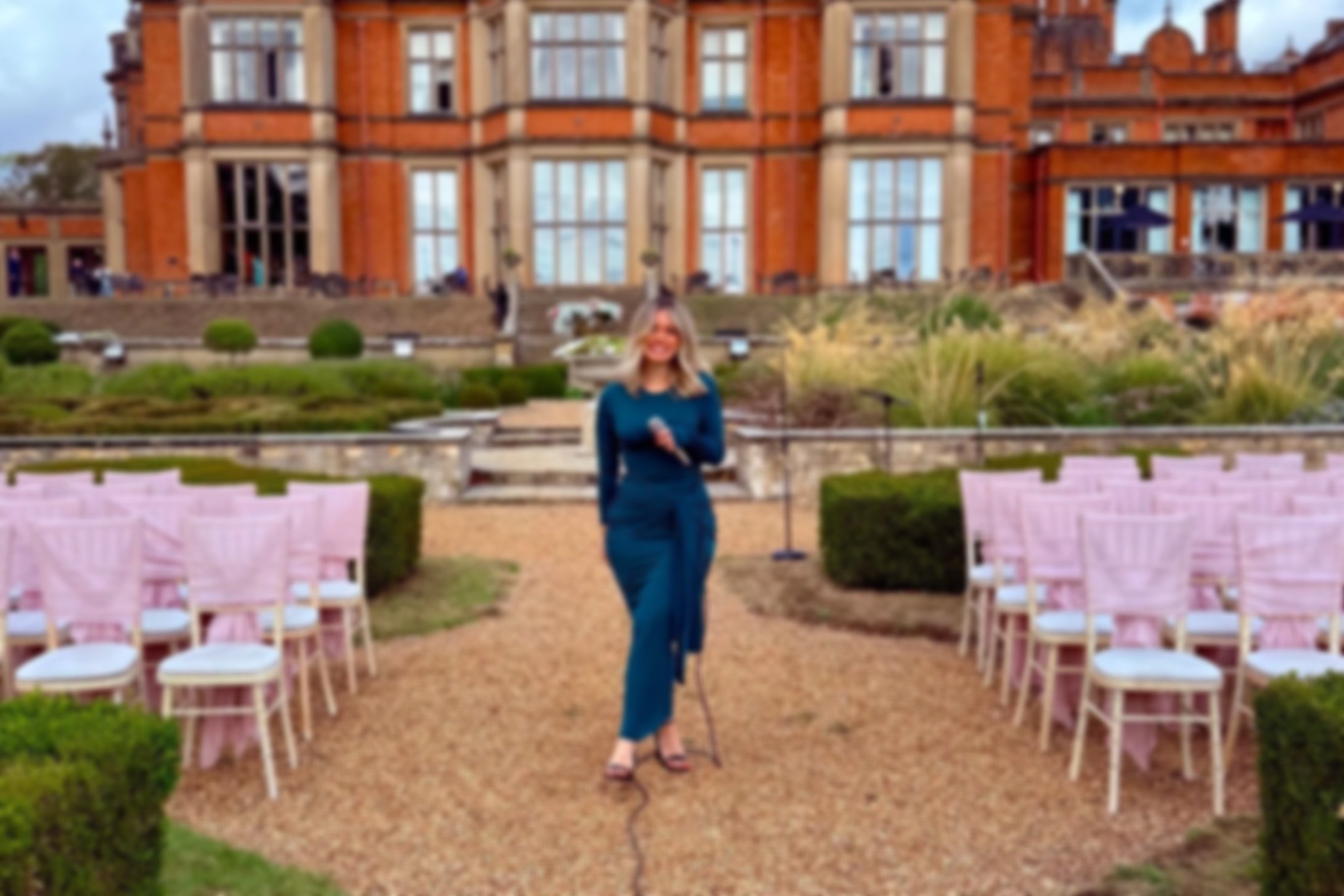 A woman standing with a microphone in hand in an outdoor wedding setup with pink chairs arranged on either side of a gravel aisle, with a large brick building in the background.