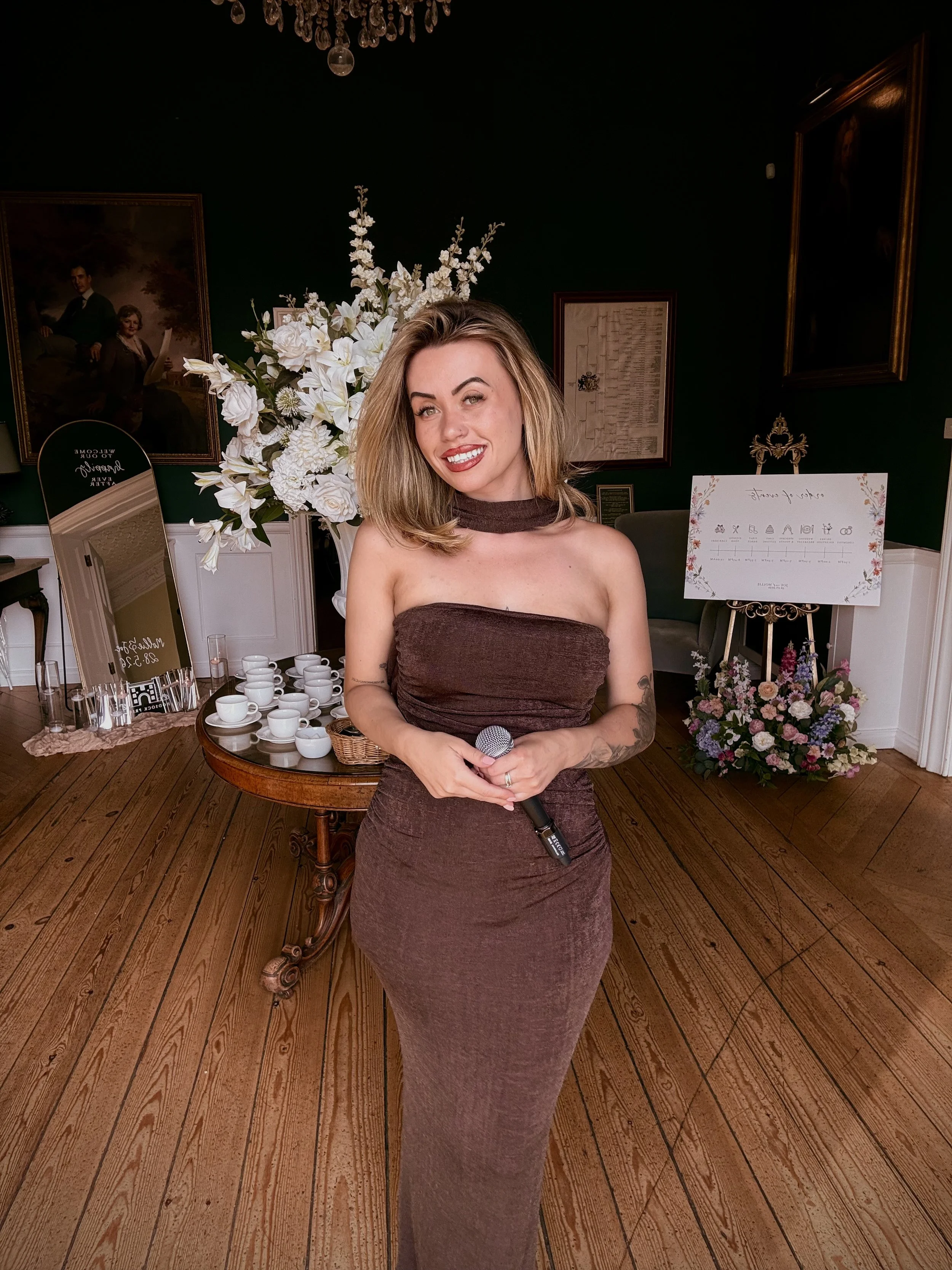 A young woman with shoulder-length blonde hair smiling while holding a microphone, standing indoors near floral arrangements and artwork.