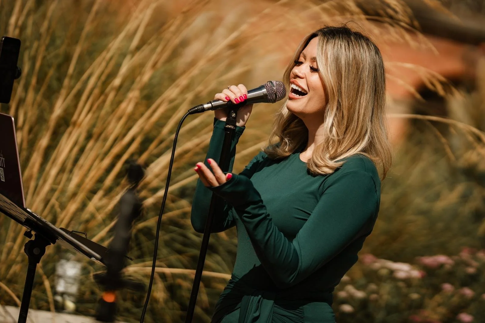 A woman singing into a vintage microphone, smiling, with framed landscape photographs on the wall behind her.