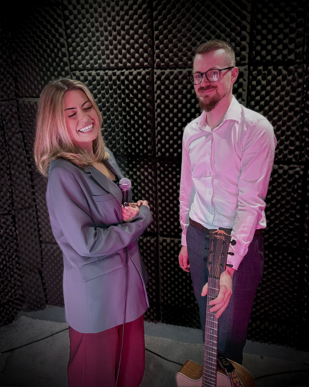 A woman holding a microphone, smiling and laughing, standing next to a man with a guitar, in a soundproof room.