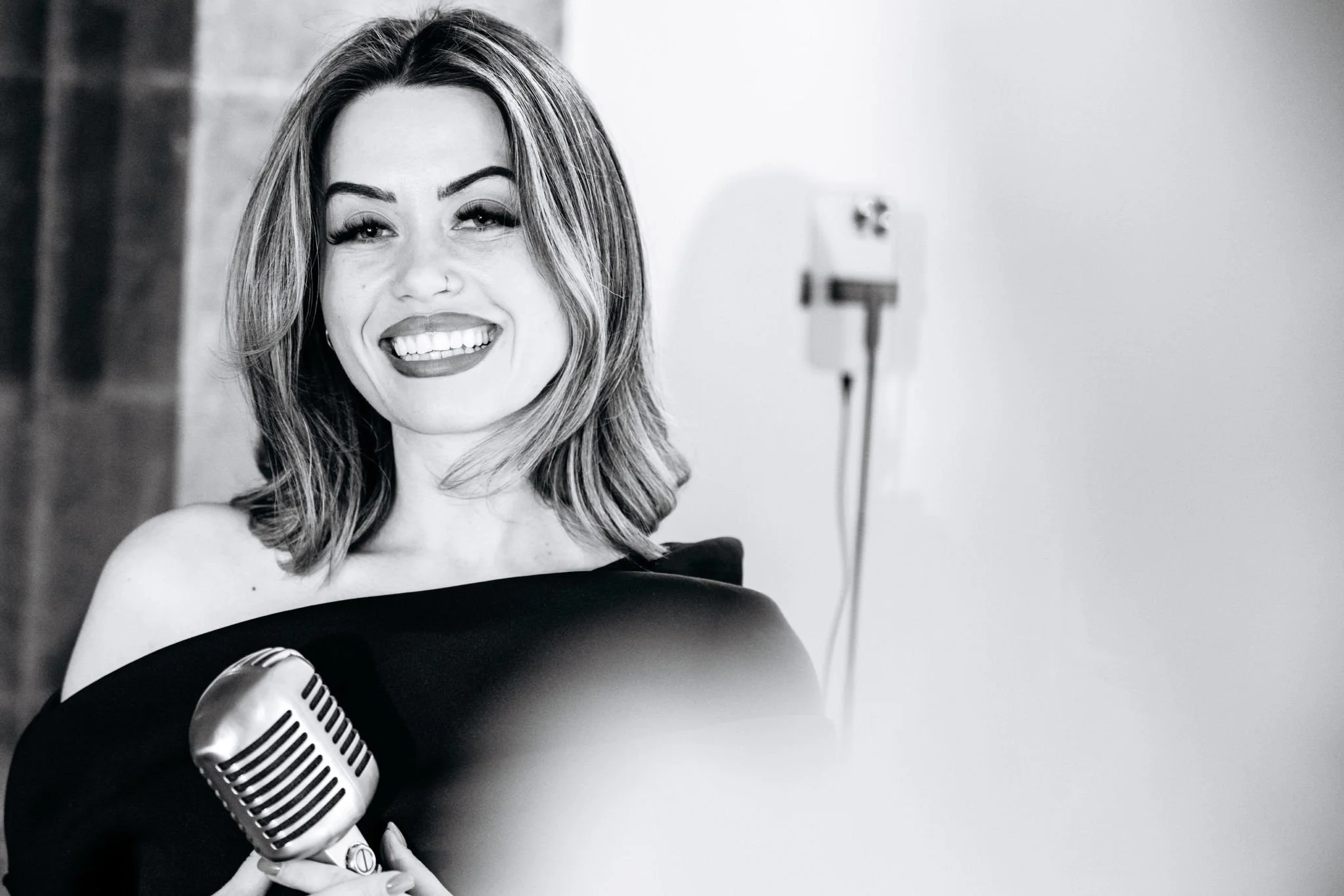 Black and white photo of a woman with shoulder-length hair, smiling, holding a vintage-style microphone, wearing an off-the-shoulder dress, in a room with plain wall and visible corded device in the background.