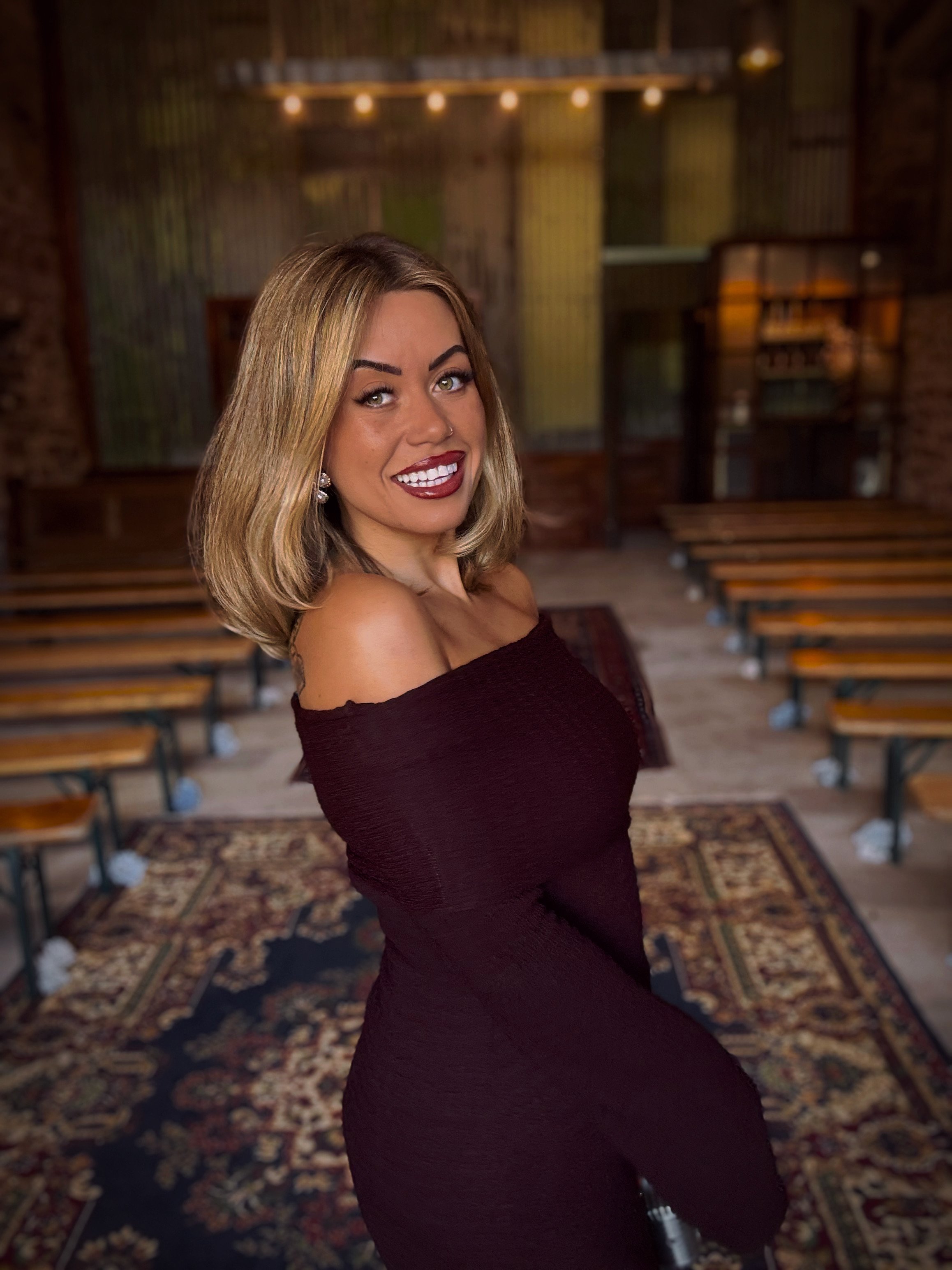 A woman with shoulder-length blonde hair, red lipstick, and earrings is smiling and looking at the camera in an indoor setting with wooden benches, a patterned rug, and a rustic background.