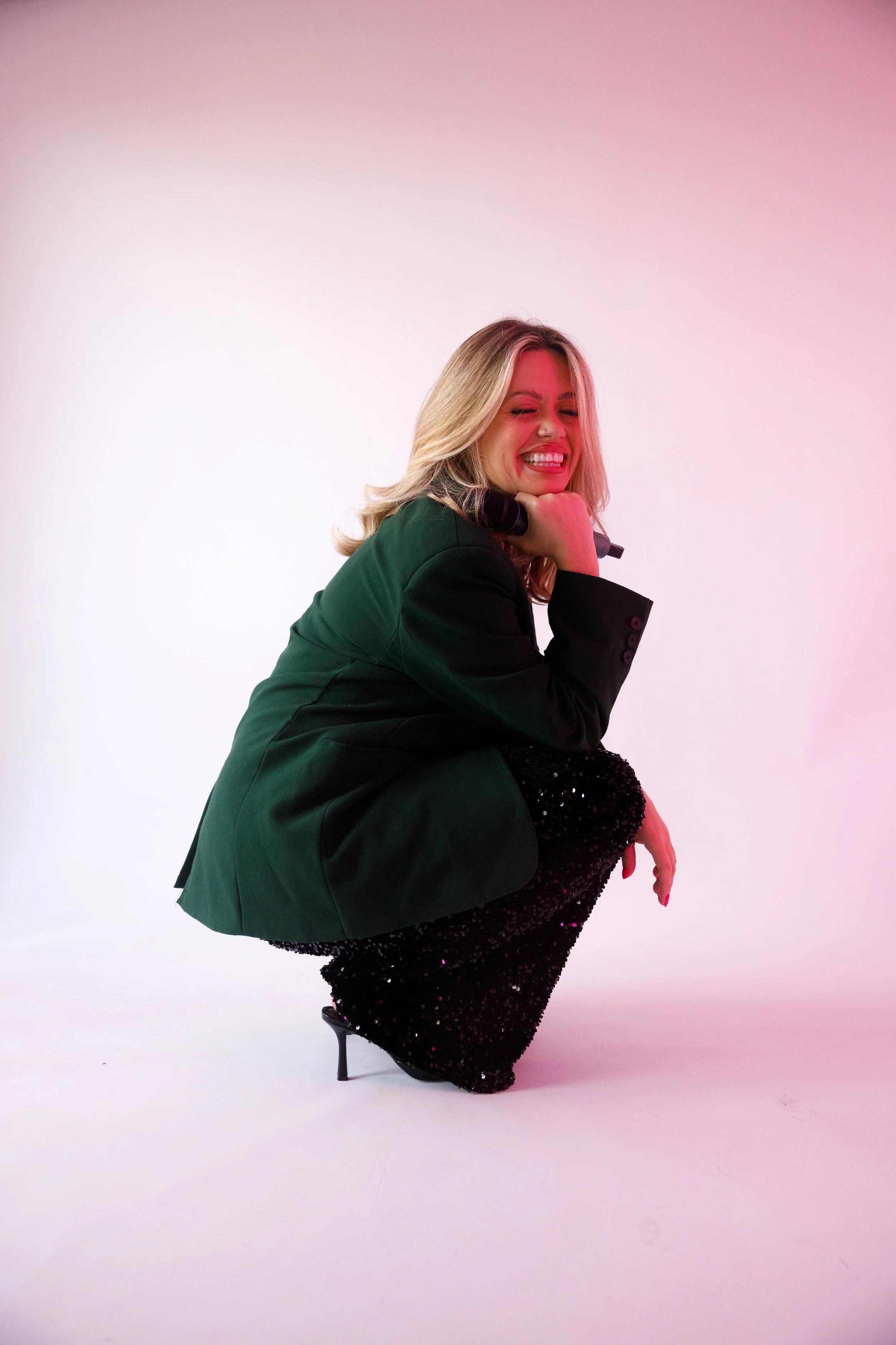 A woman with blonde hair, wearing a green blazer and a black glittery dress, is smiling and squatting in a studio with a pink gradient background.