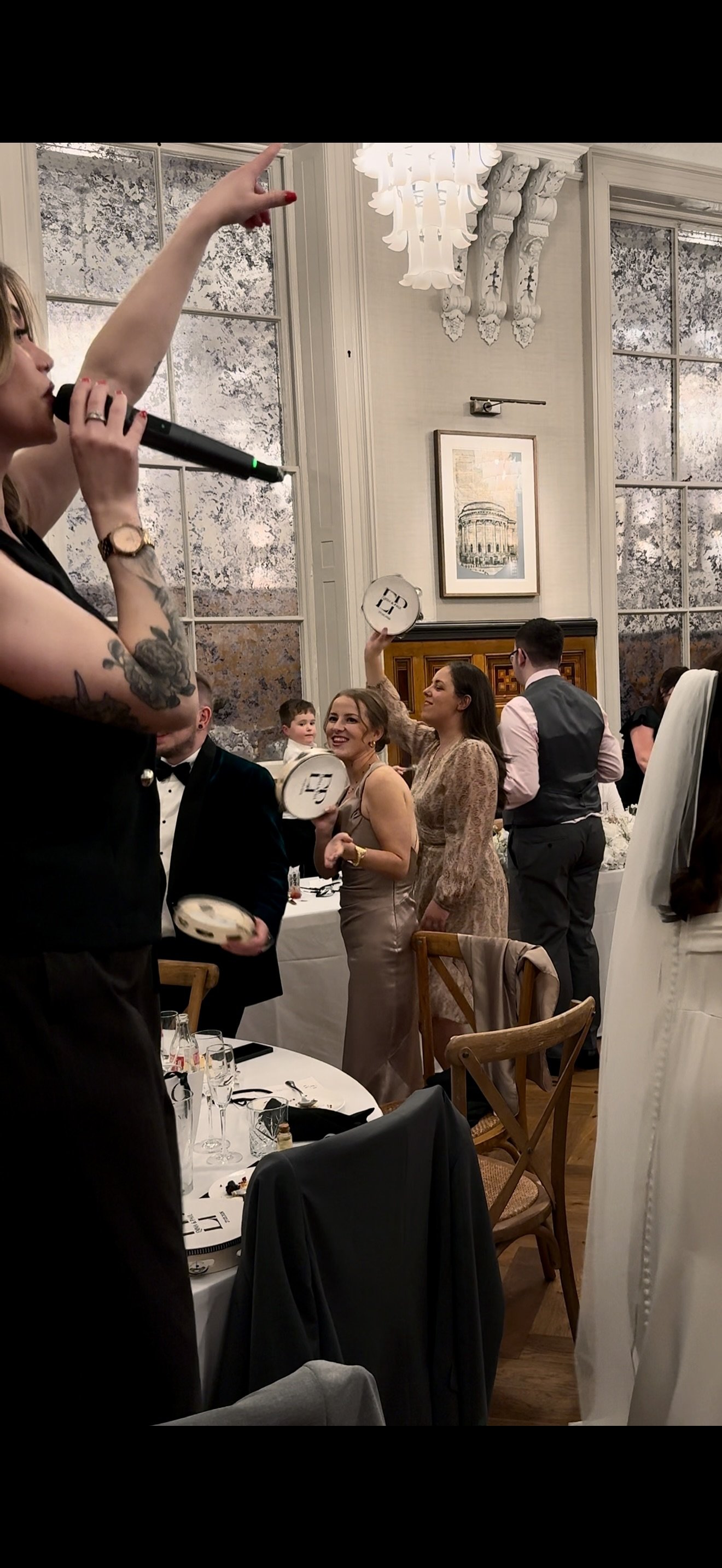 People celebrating at a formal event, some holding small plates, with a woman in a beige dress smiling and others raising their hands, in an elegant room with large windows and ornate decor.
