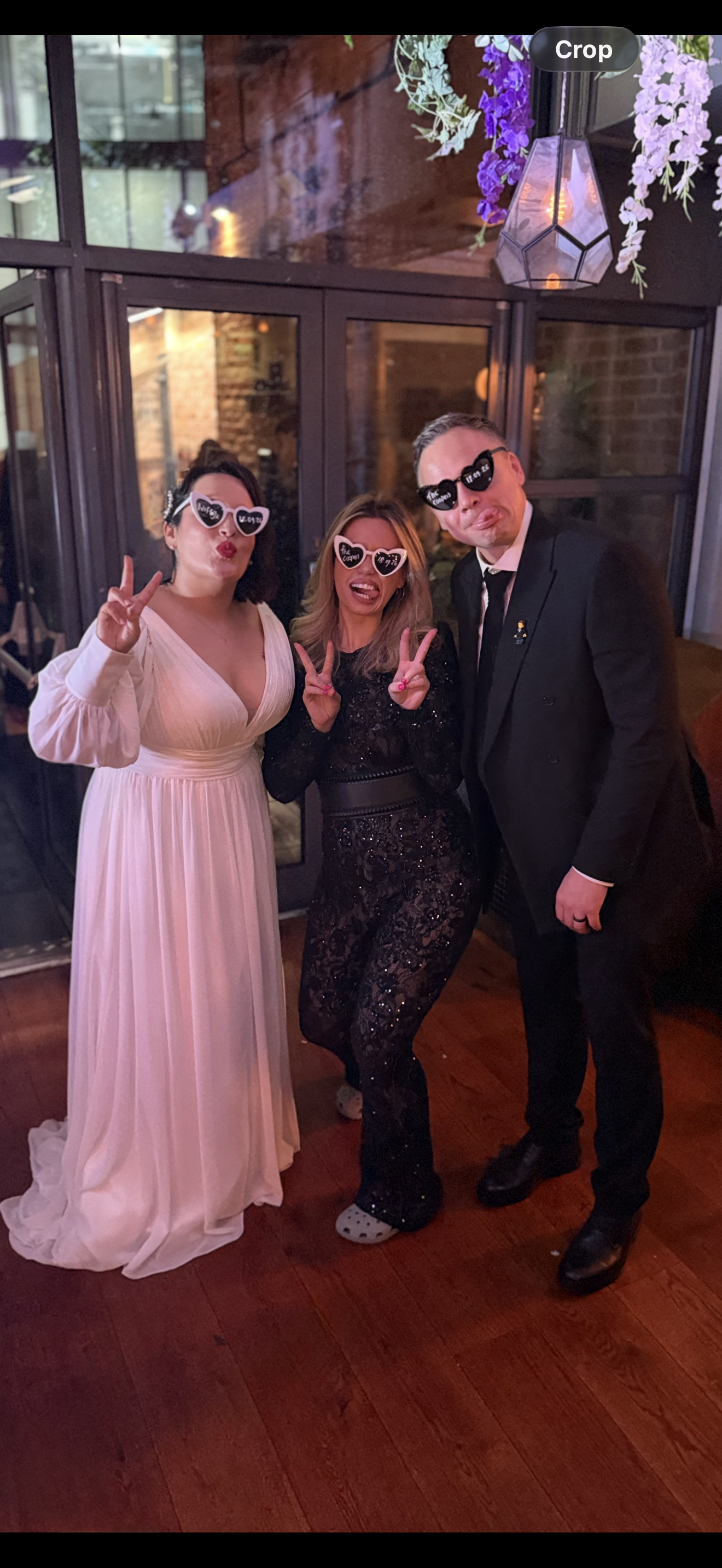 Three people at a celebration wearing heart-shaped glasses, making peace signs, and posing happily inside a venue with brick walls and wooden floors.