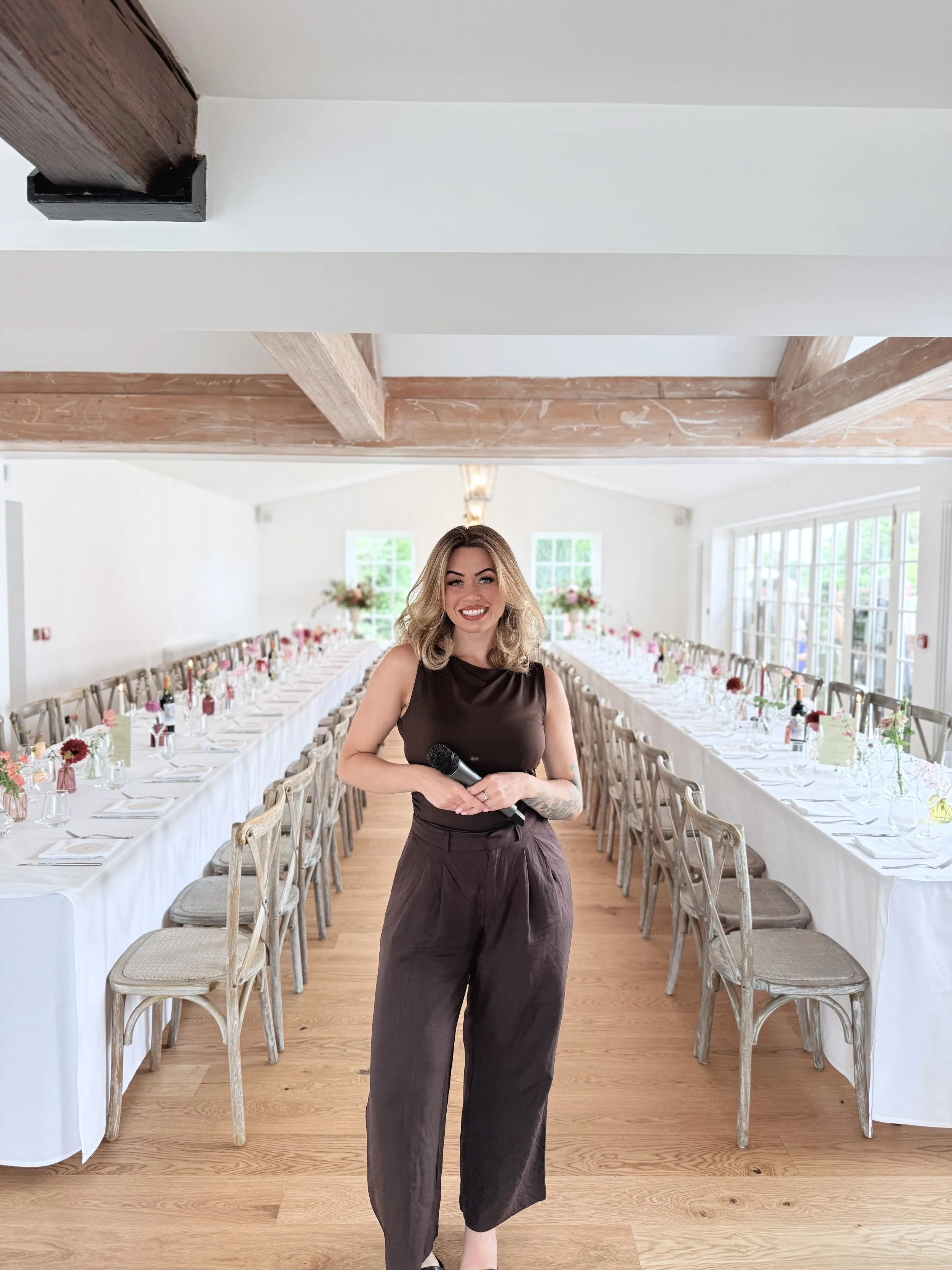 A woman with blond hair holding a microphone stands in a decorated banquet hall with long tables set for an event.